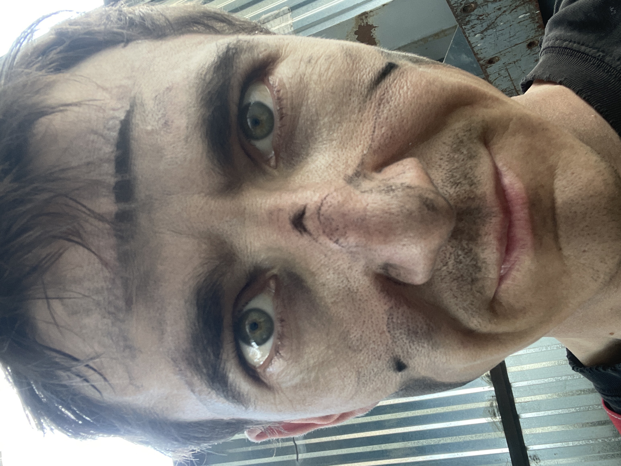 The image is a close-up, low-angle, upside-down selfie of a person's face. The subject appears to be a male with light skin, green eyes, and dark hair. His face is smudged with dirt or grease, particularly around his eyebrows, nose, and cheeks, suggesting he has been engaged in some kind of manual labor or outdoor activity. There are dark smudges that resemble war paint or decorative markings on his forehead, nose, and chin. He is smiling slightly, with his mouth open, and his eyes are wide and looking directly at the camera, conveying a sense of excitement or mischievousness.
The background is out of focus but appears to be an industrial or outdoor setting. There are corrugated metal sheets visible, suggesting a wall or structure. The lighting indicates it is daytime, possibly bright sunlight filtering in, as suggested by the bright illumination on the left side of the subject's forehead. The overall mood is informal and energetic. The provided location context of Сокуры, Russia, suggests the image was taken there. There is no visible text in the image.