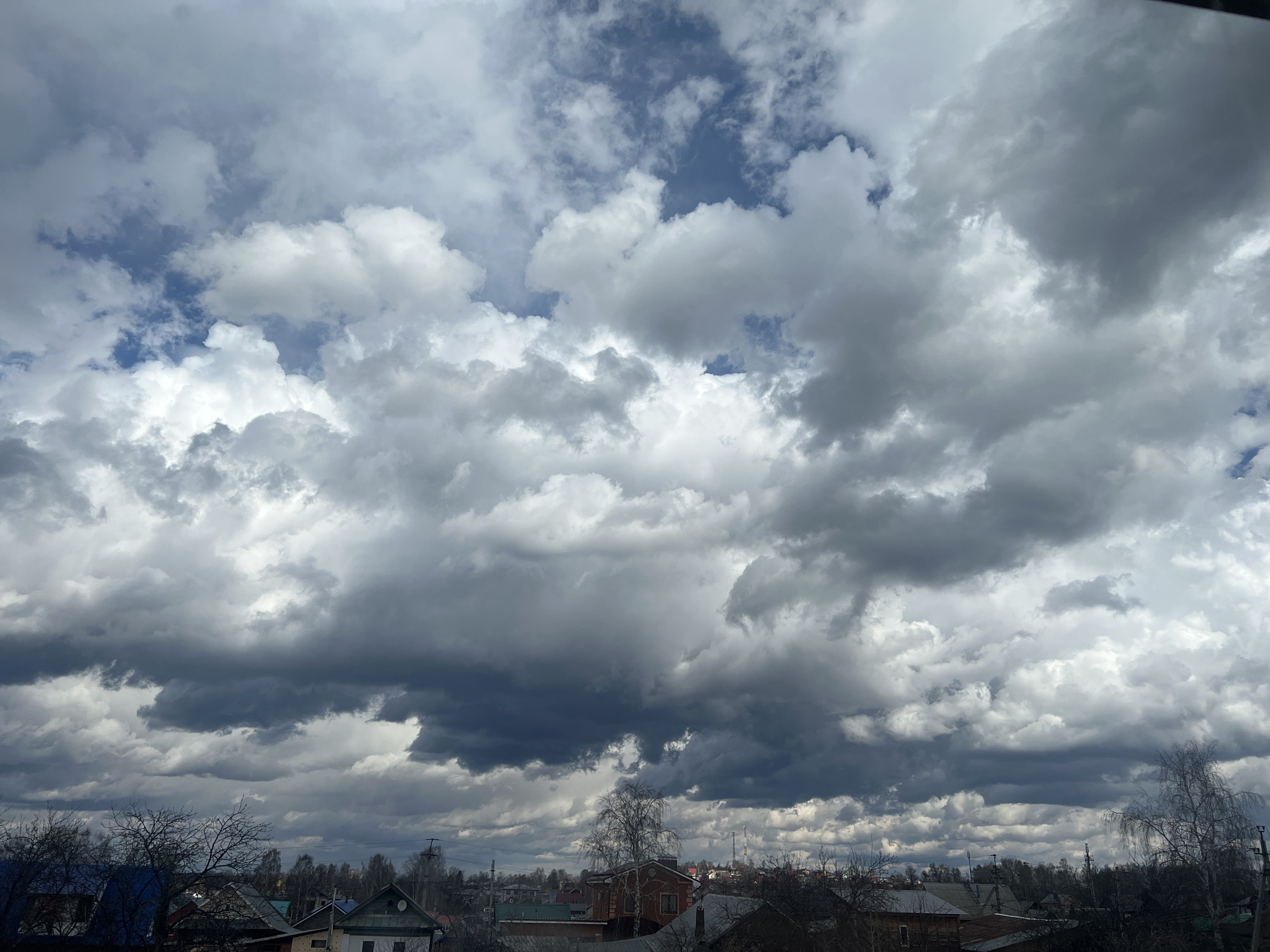 The image captures a wide view of a rural or semi-rural landscape dominated by an expansive and dramatic cloudy sky. The sky is filled with a mixture of bright white, light grey, and dark, heavy storm clouds, suggesting dynamic and unsettled weather conditions, possibly indicating an impending rain shower. Patches of clear blue sky are visible in the upper sections, contrasting with the dense cloud cover.
Below the prominent sky, the rooftops and upper stories of numerous houses are visible, extending across the lower third of the frame. The buildings display varied architectural styles, some appearing older with wooden construction, and feature roofs in different colors, including blue, green, and muted brown/grey. The landscape is dotted with numerous deciduous trees, most of which are bare, indicating late winter or early spring. A distinctive white-barked tree, possibly a birch, stands out in the mid-ground. Utility poles and power lines are discernible among the trees and houses. In the distant background, faint structures resembling communication towers can be seen on a raised elevation.
The scene depicts a quiet daytime environment in Votkinsk, Russia. No people or specific activities are visible, lending a calm, contemplative, and slightly moody atmosphere to the image due to the heavy clouds. There is no visible text in the media file.