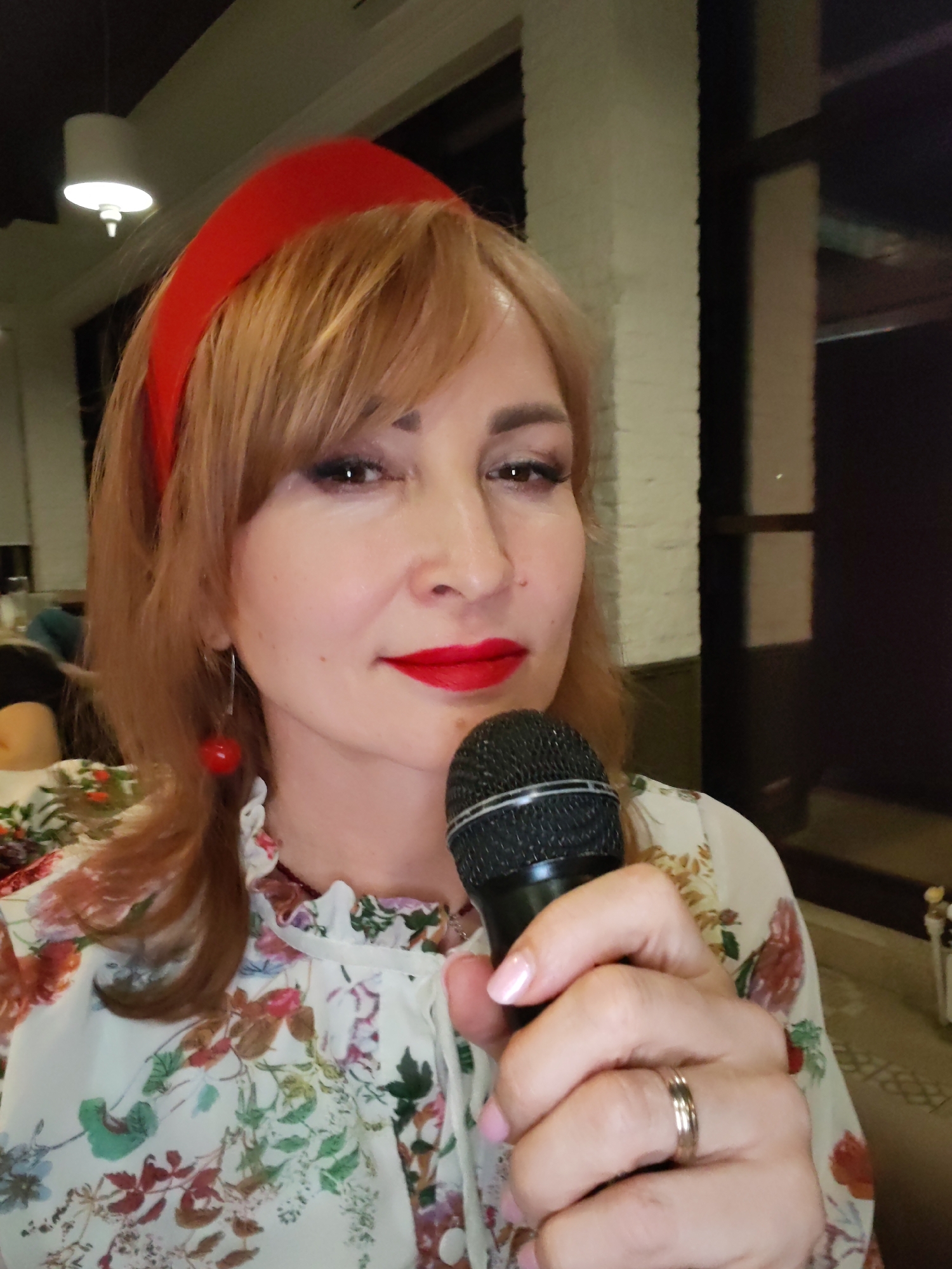 A woman is holding a microphone and facing the camera. She has red lipstick, wears a red headband, and has red cherry earrings. She is wearing a white floral shirt. On her ring finger is a ring. The location appears to be indoors, near a window and some interior lighting. There is no indication of the city of Chaikovsky, Russia, in the image.