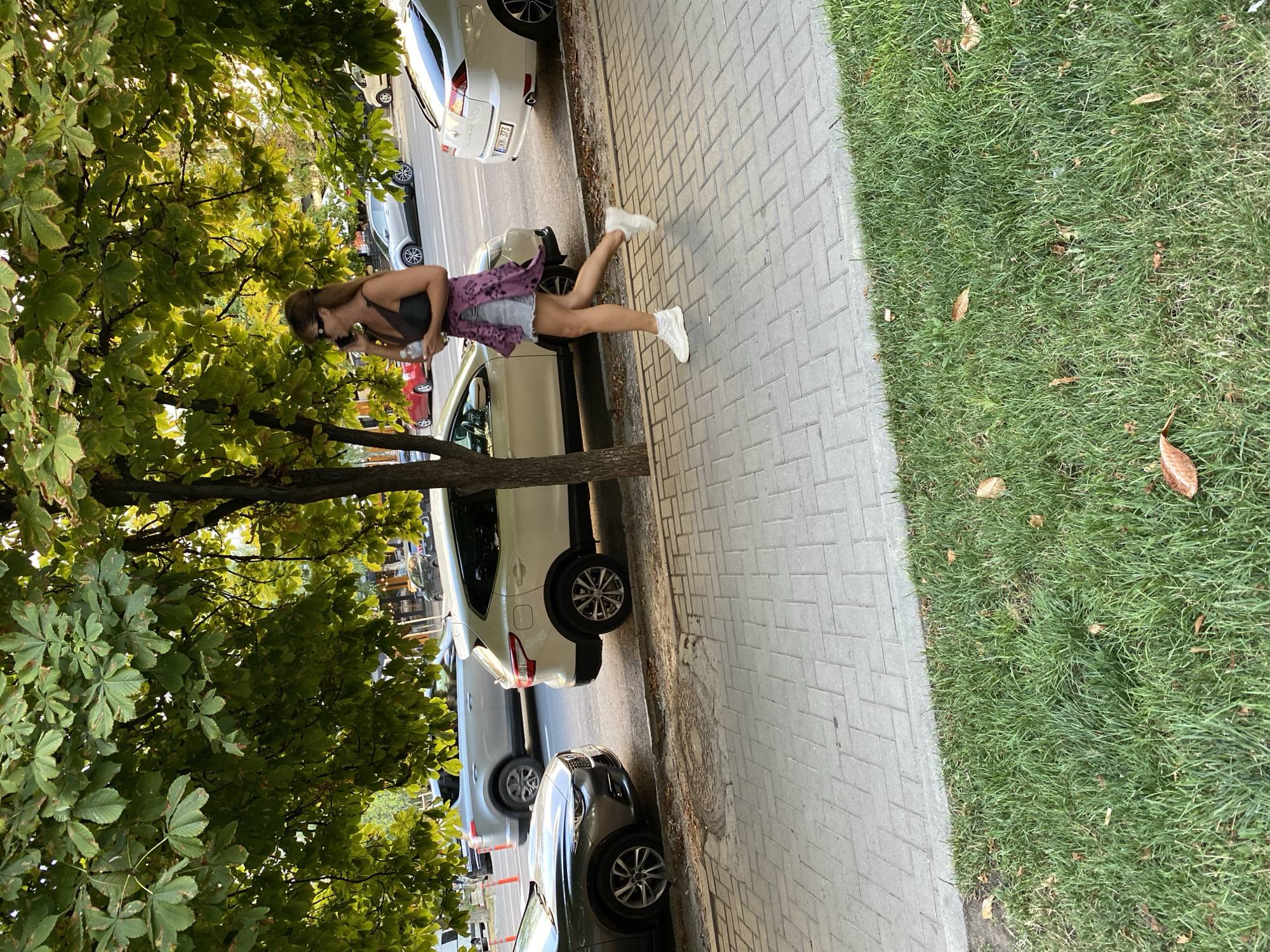 In Chișinău, Moldova, a woman is walking on a sidewalk next to a street lined with parked cars and trees. She is wearing a dark tank top, denim shorts, a purple open shirt tied around her waist, and white sneakers. She is holding a clear bottle in her left hand and talking on her phone with her right hand. The sidewalk is paved with rectangular grey bricks, and a strip of green grass runs alongside it. Several cars are parked along the street, including a white SUV, a silver sedan, and a darker colored sedan further down. The trees overhead cast dappled shade on the scene.