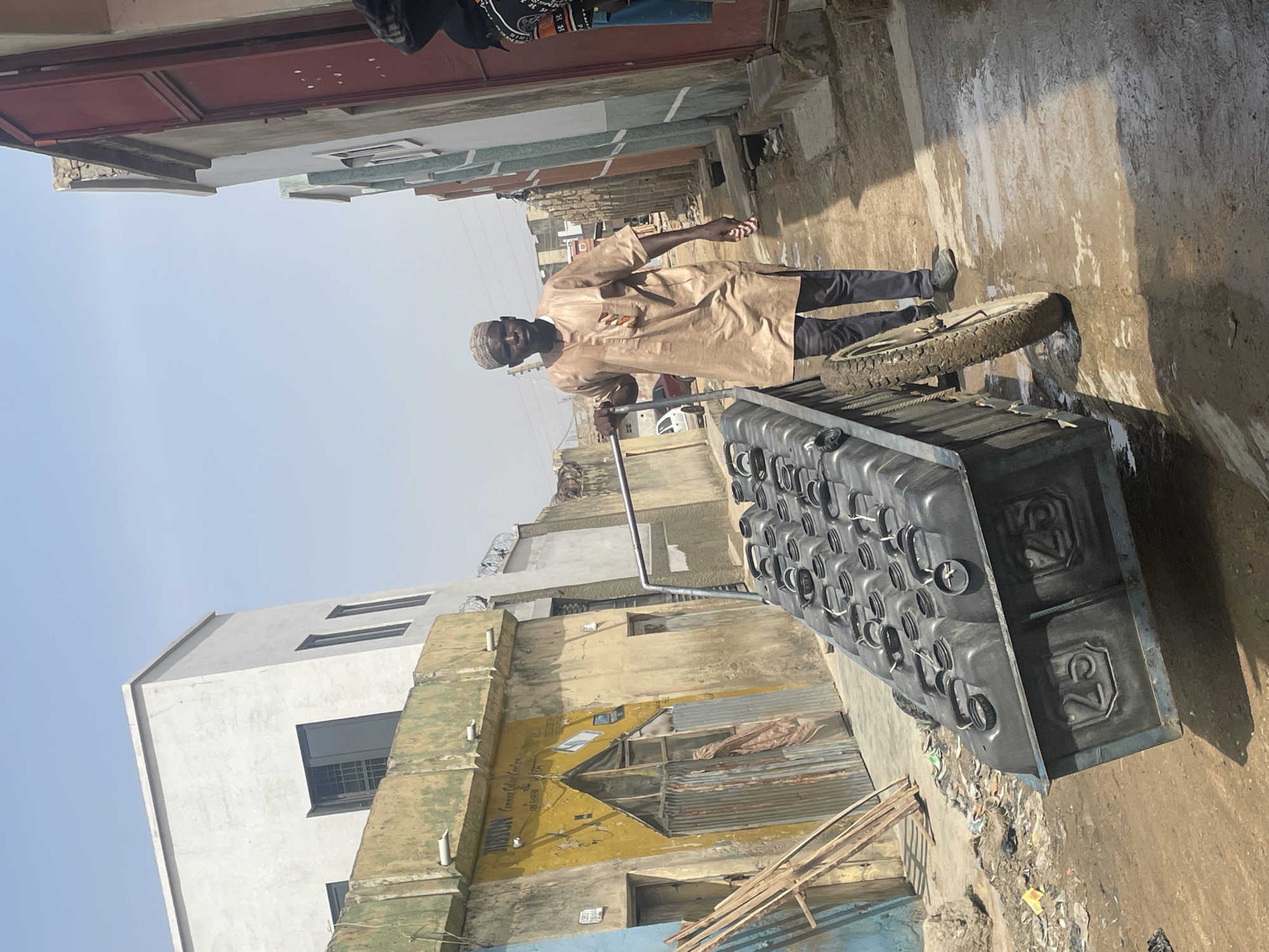 In a street scene, an adult male wearing a tan robe and dark trousers is pushing a large, heavily loaded cart through a somewhat narrow alleyway. The cart is packed with numerous black containers, possibly jerrycans, stacked uniformly. The ground appears damp and possibly muddy. The man has a dark complexion and is wearing what looks like a small, patterned cap.
The environment consists of buildings on both sides of the alley, showing signs of age and wear. The buildings have a mix of paint colors, including yellow, white, and a touch of blue. There's what appears to be a mural or design on one of the buildings. The sky is visible in the background and appears pale blue or hazy.
The location appears to be an urban residential area, based on the type of buildings and alleyway. The exact city is unknown, and location "Unknown, Unknown" is given.