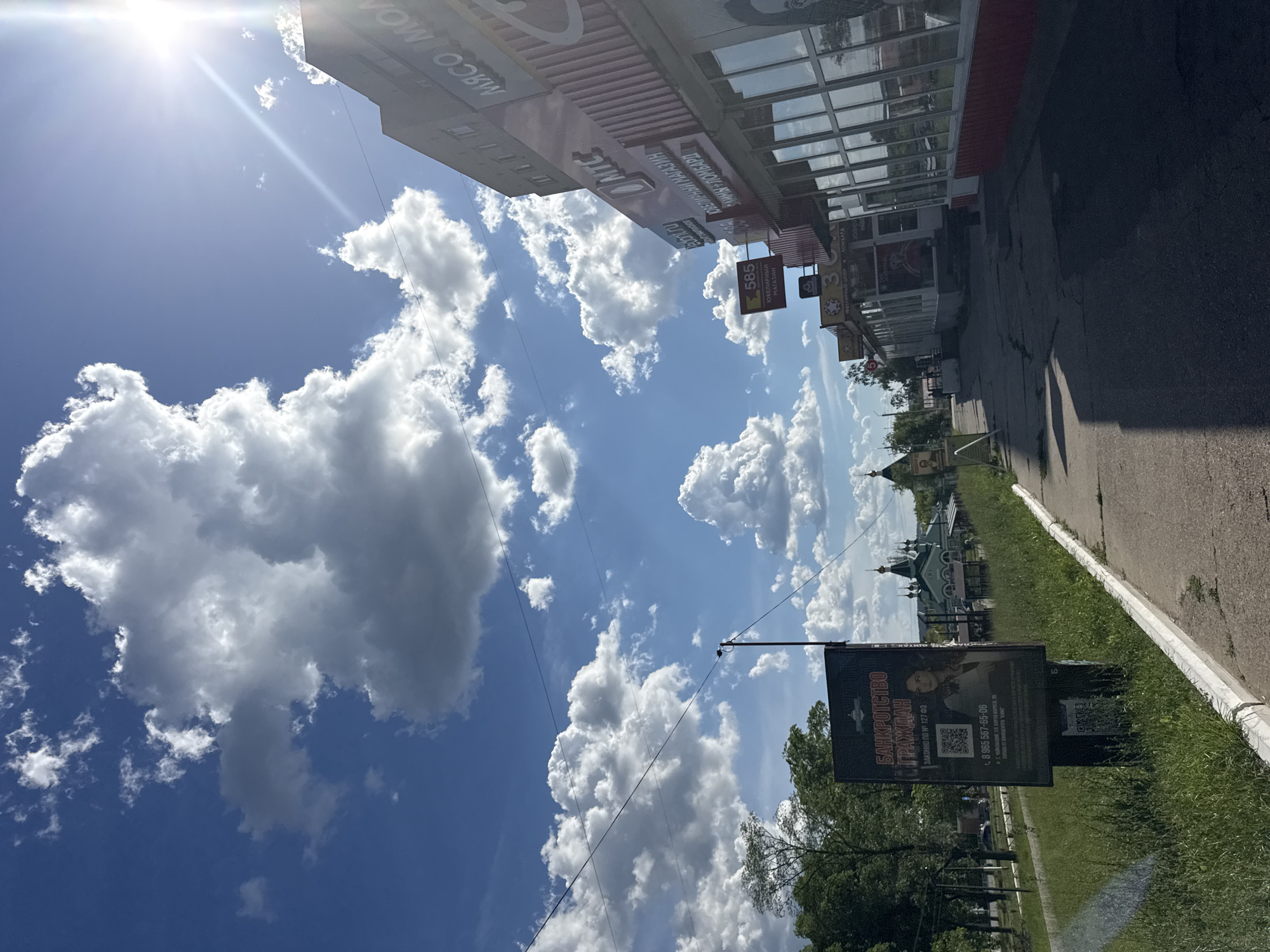 This is a vertical, eye-level, outdoor photograph taken on a sunny day with scattered white clouds in a bright blue sky. The scene appears to be a street in Chaikovsky, Russia.
In the foreground, on the right side of the image, is a paved sidewalk and a grassy area. A black billboard is prominently displayed, advertising "BANKRUPTCY OF CITIZENS" and providing a phone number, website, and a QR code. The text on the billboard is in Russian. To the left of the billboard, there is a street with some trees and a building with a red roof. Further down the street, a church with green domes is visible.
The left side of the image features the side of a multi-story building with large windows and a red awning. The sun is shining brightly in the upper left corner, creating a lens flare. Signs on the building are partially visible and also in Russian, including "585 JEWELRY STORE." The overall impression is a sunny day in a Russian town, with commercial establishments lining the street. The image appears to have been taken from a moving vehicle or by someone walking along the street.