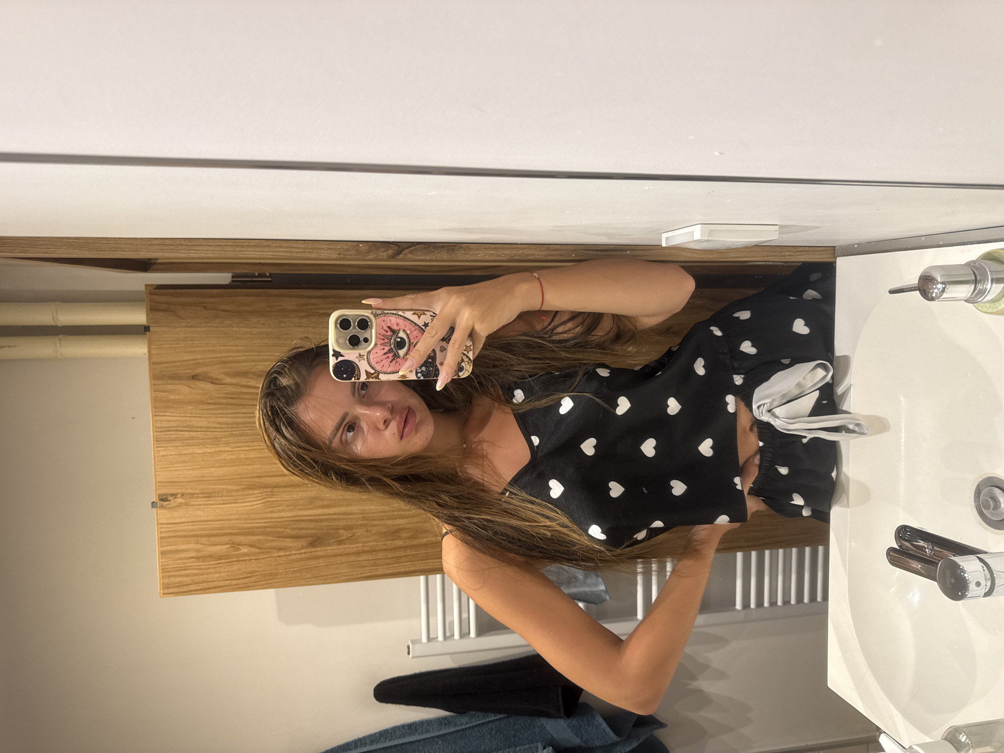 A young woman with long, wet brown hair is taking a selfie in a bathroom mirror. She is wearing a black pajama set with white hearts. Her right hand is on her hip, and she is holding a phone in her left hand. The phone case is pink with an eye design. The bathroom has a white sink with silver faucets, a wooden cabinet, and a towel rack with towels. The image is taken in a residential bathroom.