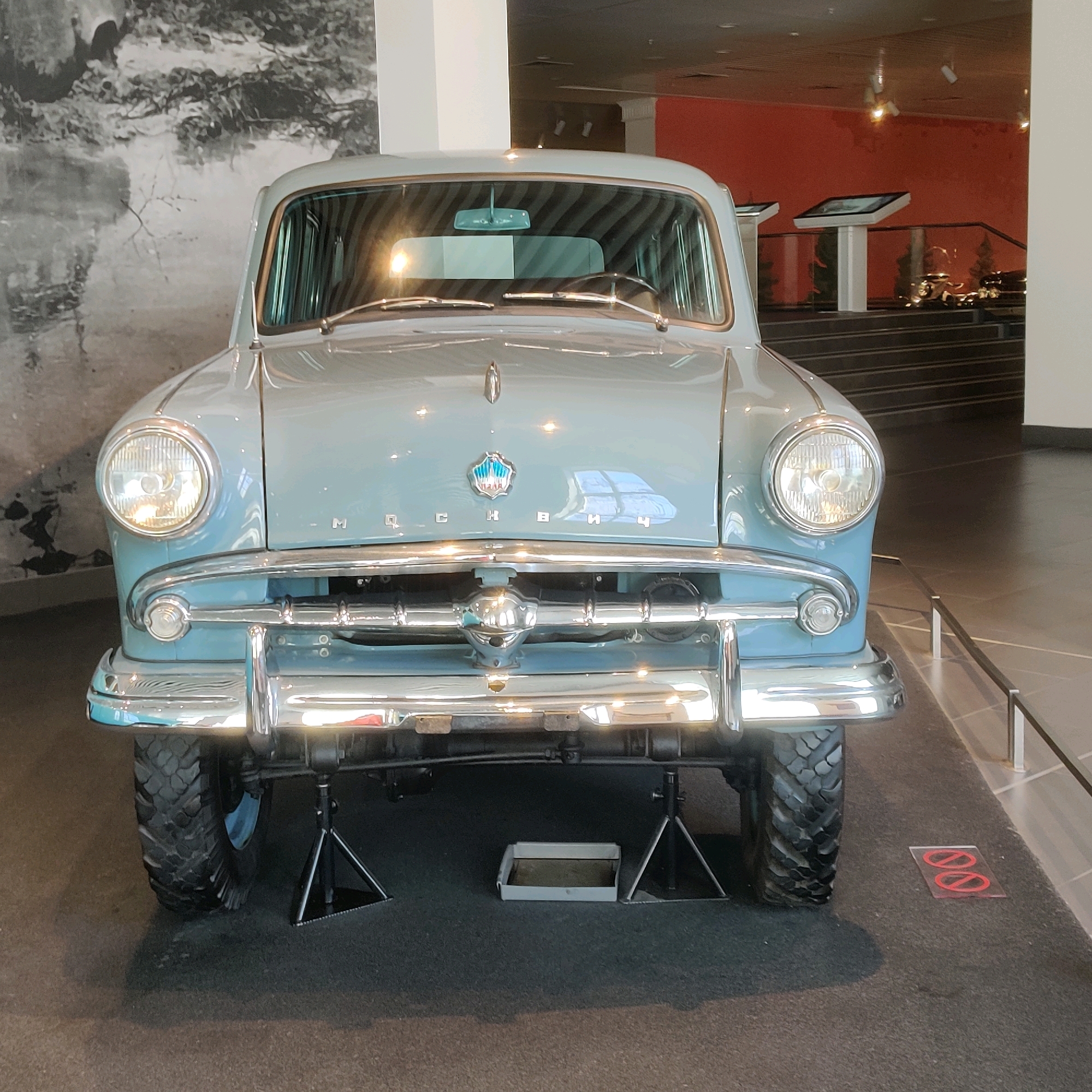 This is a front-facing, eye-level photograph of a light blue vintage car on display indoors. The car is a Moskvitch, identifiable by the chrome emblem and lettering on the hood. It is elevated on jack stands, revealing its underside and a drip pan underneath. The car has large, round headlights and a prominent chrome bumper. The background shows a museum-like setting with a large black and white mural on the left wall, and a red-colored wall with display screens and stairs on the right. The floor is dark grey carpet. This scene is located in the Museum of Military Technology "Battle Glory of Urals" in Verkhnyaya Pyshma, Russia.
