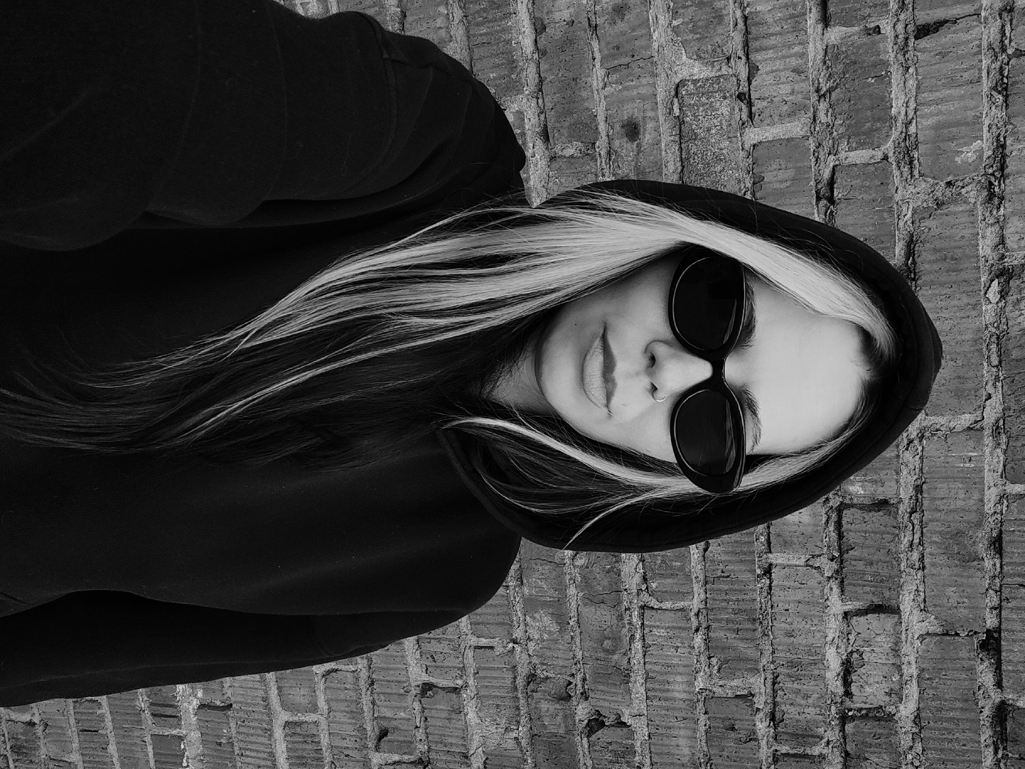 A grayscale, angled close-up depicts a person wearing a dark hooded sweatshirt and oval sunglasses. The person has long hair with blonde highlights. The background is a brick wall. Location data is not ascertainable from the image.