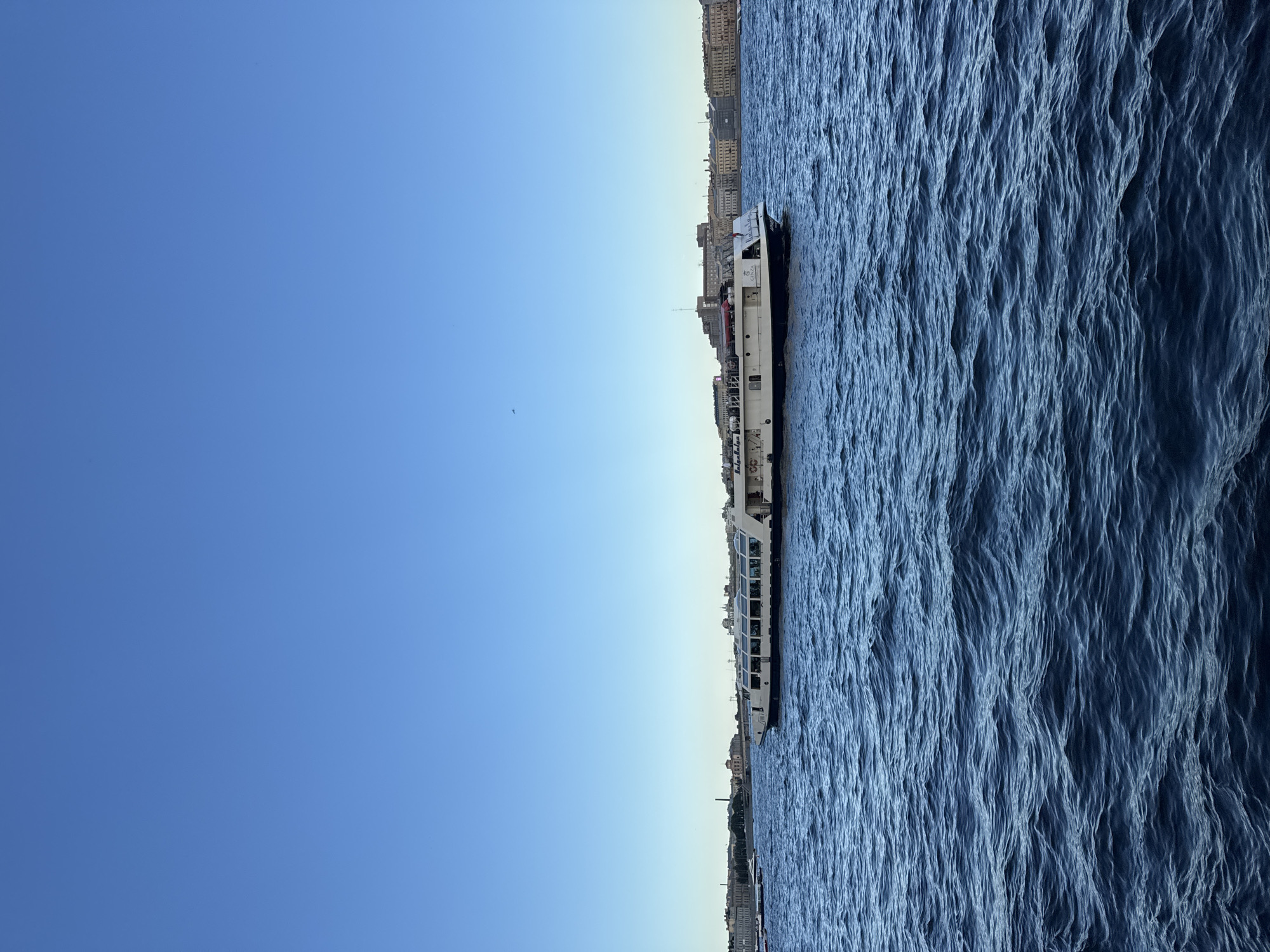 A wide shot captures a long, white and black passenger boat navigating across a deep blue, choppy river under a clear, bright blue sky. The boat, which appears to be a tour boat, has multiple windows on its upper deck and a darker hull. In the distance, a city skyline is visible with a mix of older, classical architecture and some more modern buildings. A bridge can be seen spanning the river in the background. The overall scene suggests a pleasant day in Saint Petersburg, Russia, likely during the warmer months. The water is a prominent element, filling the lower half of the image with its texture and color.
