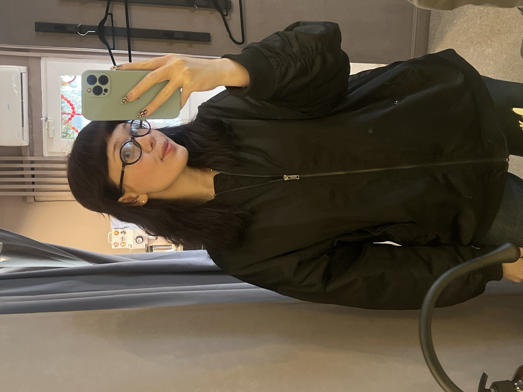 A woman with black hair and glasses takes a selfie indoors. She is wearing a black bomber jacket. The background features a window, a radiator, and coat hangers. Other details include a phone case and a curtain. The location is within a fitting room. There is no confirmation that the location is in the city of Chaikovsky, Russia.