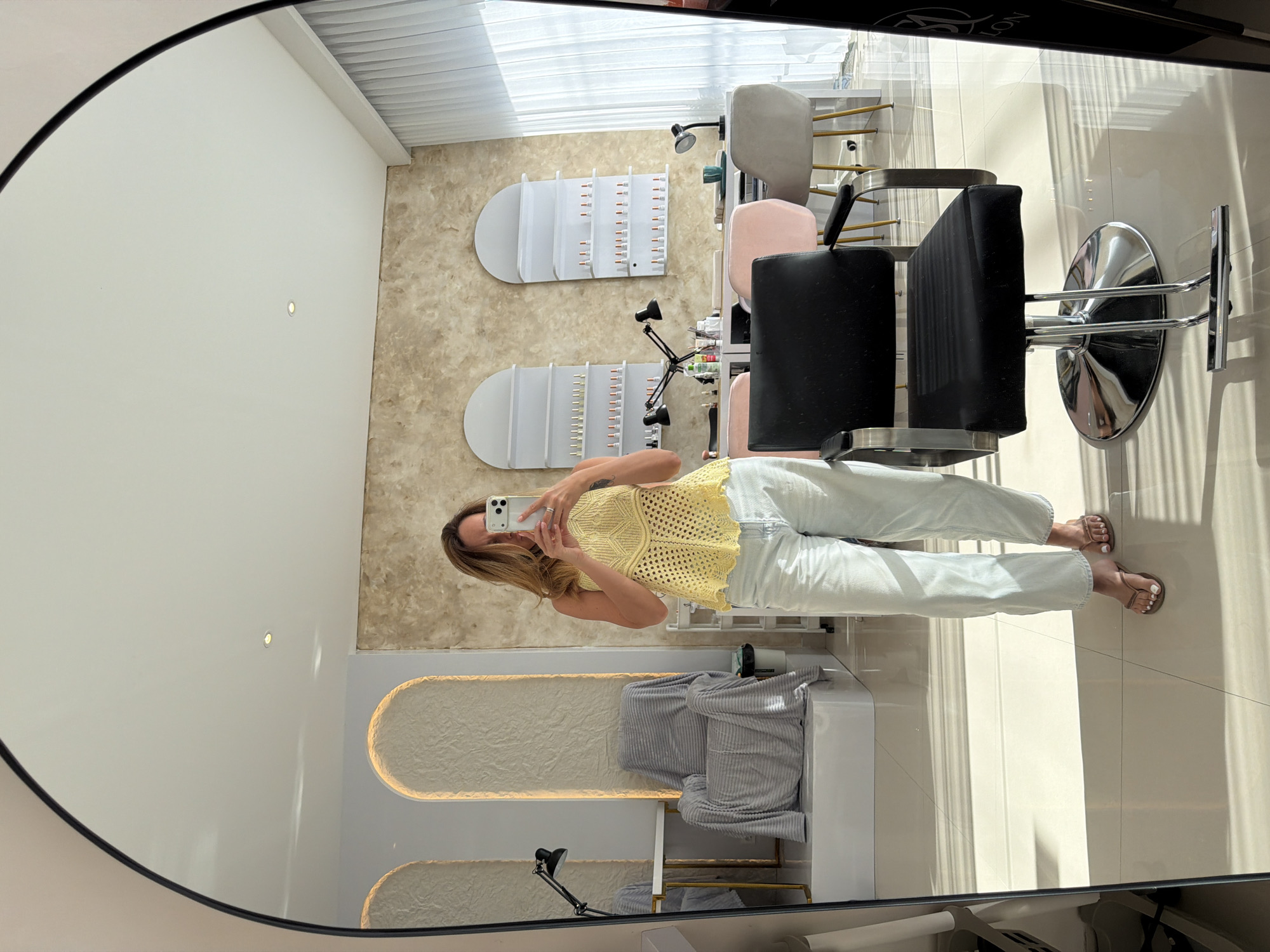 The image is a reflection in an oval mirror, likely in a commercial setting, possibly a beauty salon or nail spa in North Kuta, Indonesia. A woman is visible, standing in the reflection, holding her phone up to take a selfie in the mirror. She is wearing a light yellow crocheted top and light-wash jeans, along with brown sandals. The room appears modern, with white walls and flooring. There are several manicure stations. Each station has a semi-circular, illuminated back panel, and a nail drying device. There are also salon chairs with shiny metal bases. The lighting seems to be a mix of natural and artificial light, with sunlight streaming in from a window, as well as overhead lights. Overall, the scene is clean, well-lit, and appears to be a commercial beauty salon.