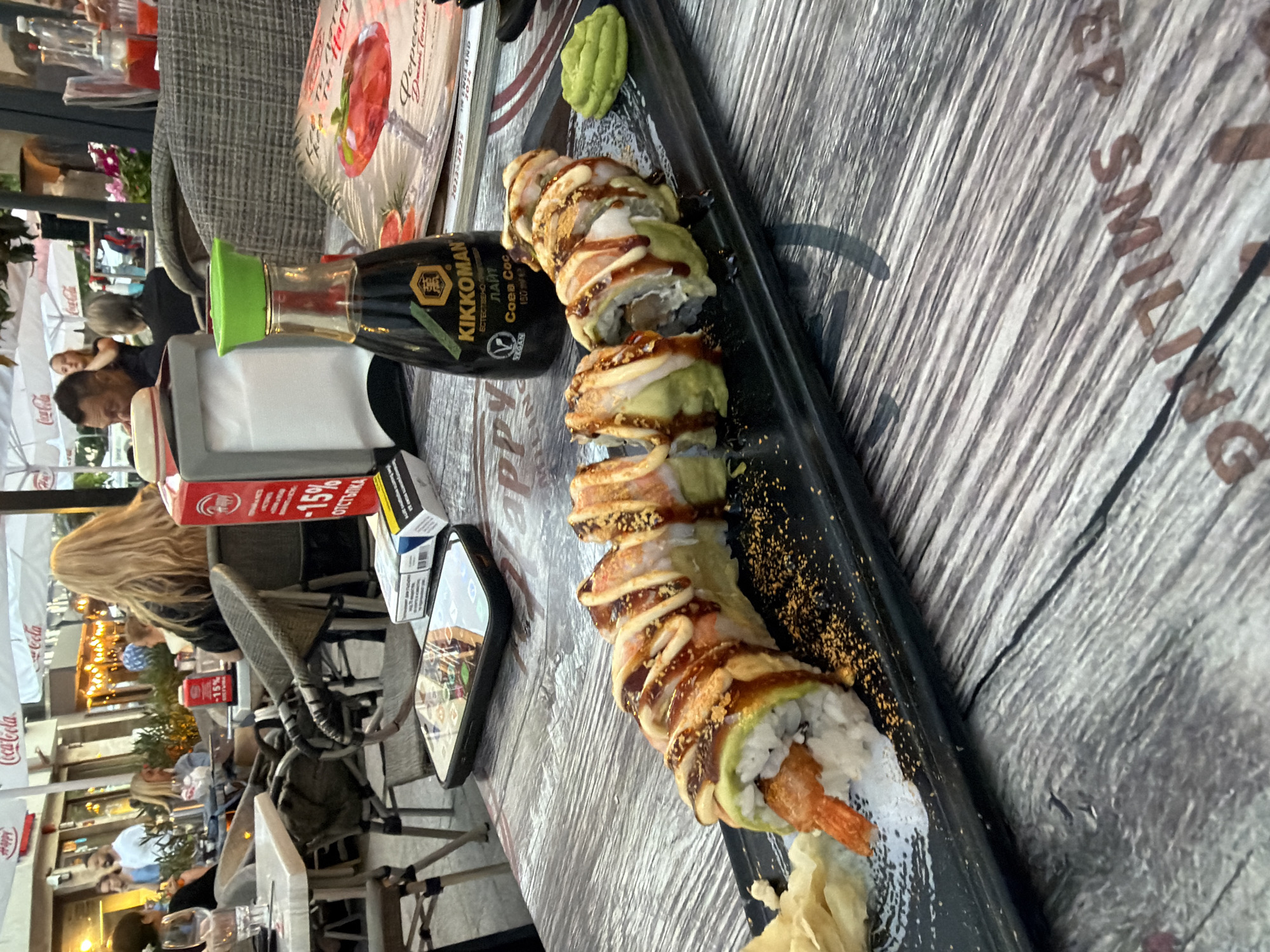 A close-up shot shows a plate of sushi and various items on a table at an outdoor restaurant in Burgas, Bulgaria. The sushi is served on a black rectangular plate, accompanied by pickled ginger and a dollop of wasabi. A bottle of Kikkoman soy sauce stands behind the sushi. A smartphone is visible in the foreground, resting on the table. In the background, patrons are seated at tables under umbrellas. The scene appears to be an evening setting, with warm lighting illuminating the area. The restaurant's proximity to the sea in Burgas is suggested by the outdoor seating and the casual ambiance.