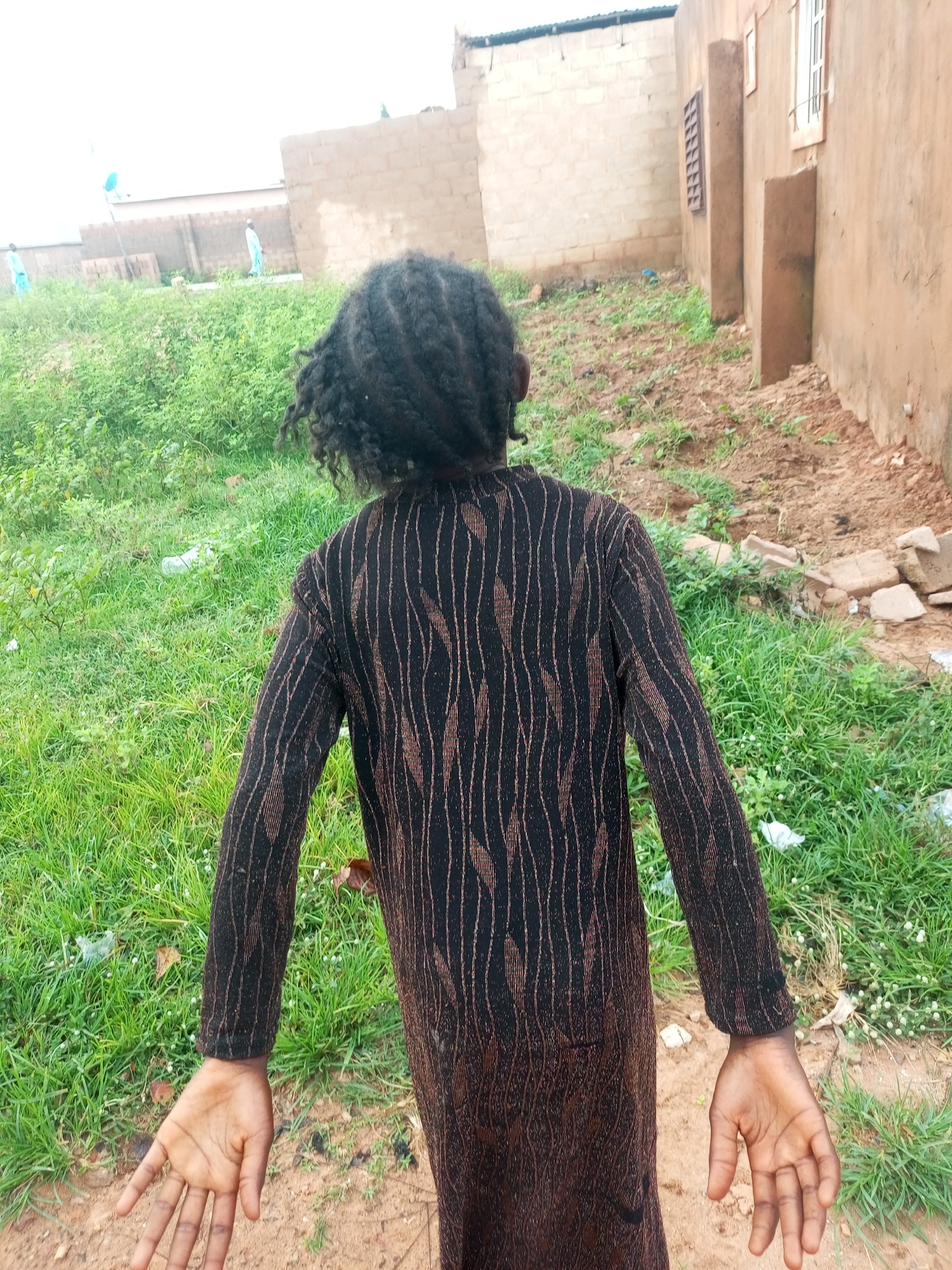 An overhead rear view shows a child with dark, braided hair and wearing a dark long-sleeved garment with an all-over vertical pattern of thin, light brown or copper-colored lines, standing in a patch of dry, reddish-brown dirt with sparse green weeds and debris. The child's arms are outstretched to the sides, palms facing upward. Behind the child, a slope of overgrown green grass and weeds stretches towards a beige, brick-building in the background. To the right, the side of a rough, sandy-colored building with a single window opening is visible. The environment suggests an outdoor area in a rural or semi-urban setting, possibly a residential area with unfinished construction, in Tempure, Nigeria.