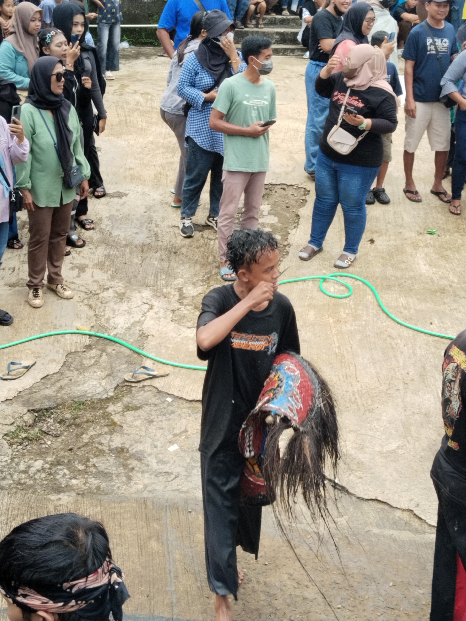 In Kuto, Indonesia, an elevated, open-air concrete platform is populated with a mixed-gender crowd of adults, many of whom are masked and/or holding or wearing head coverings (hijab, headbands). The people stand and observe. The foreground is dominated by a young man holding a headdress or mask decorated with red, black, blue, and white paint and long strands of dark hair or fiber. He wears a black t-shirt and wet-looking black pants. The ground around and between the people is paved with concrete. A green garden hose lies on the pavement, and several pairs of sandals rest nearby. The people in the background stand on concrete steps.