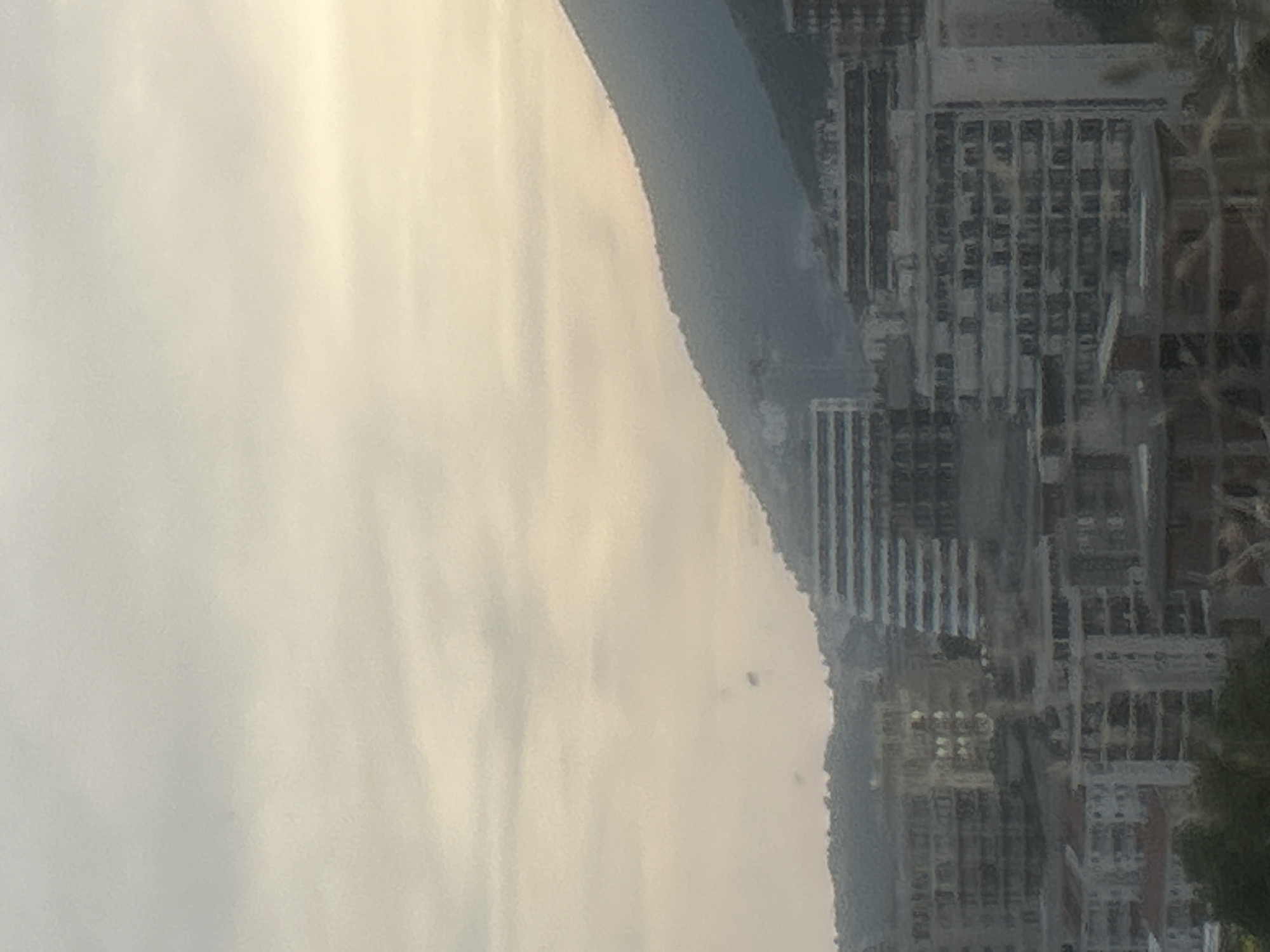 The image shows a view of the city of Rafailovići, Montenegro, with a large mountain in the background. The sky is overcast and hazy, with a pale yellow and white color palette. In the foreground, there are several buildings, some of which appear to be under construction. The buildings are multi-story and have a modern architectural style. The overall scene is somewhat muted due to the atmospheric conditions.