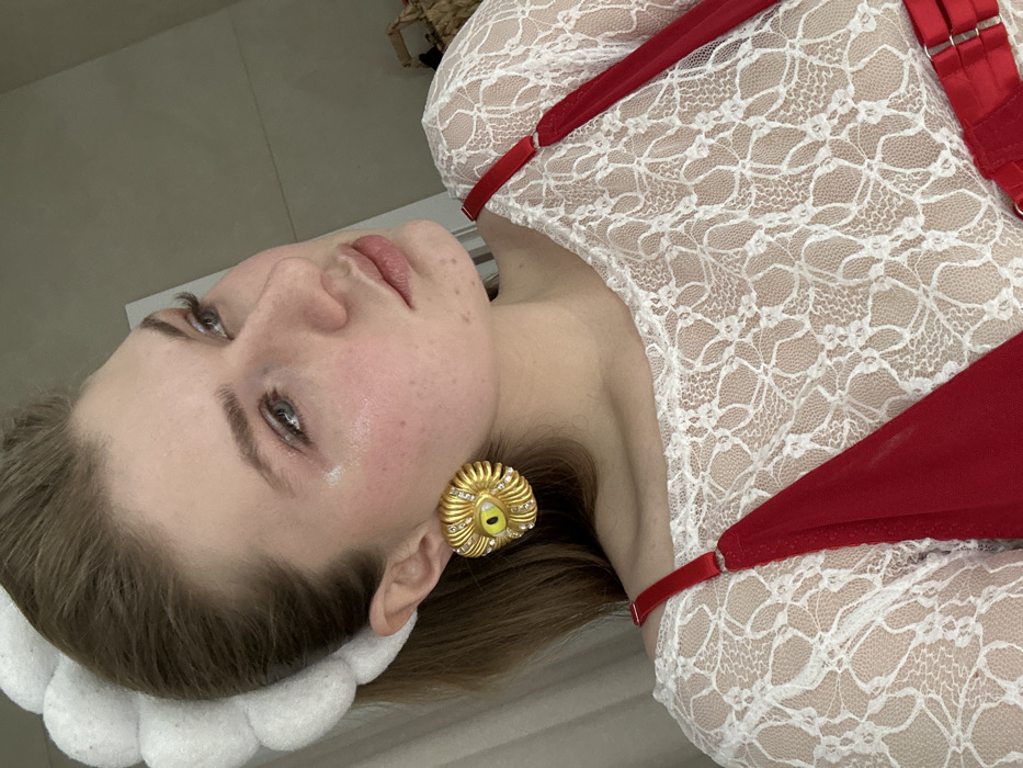 A woman with light skin and brown hair poses. She wears a white lace top with red straps. A large, gold, eye-shaped earring adorns her ear. Her head rests on a white, floral-patterned cushion or headband. The background appears to be a plain, light-colored surface.