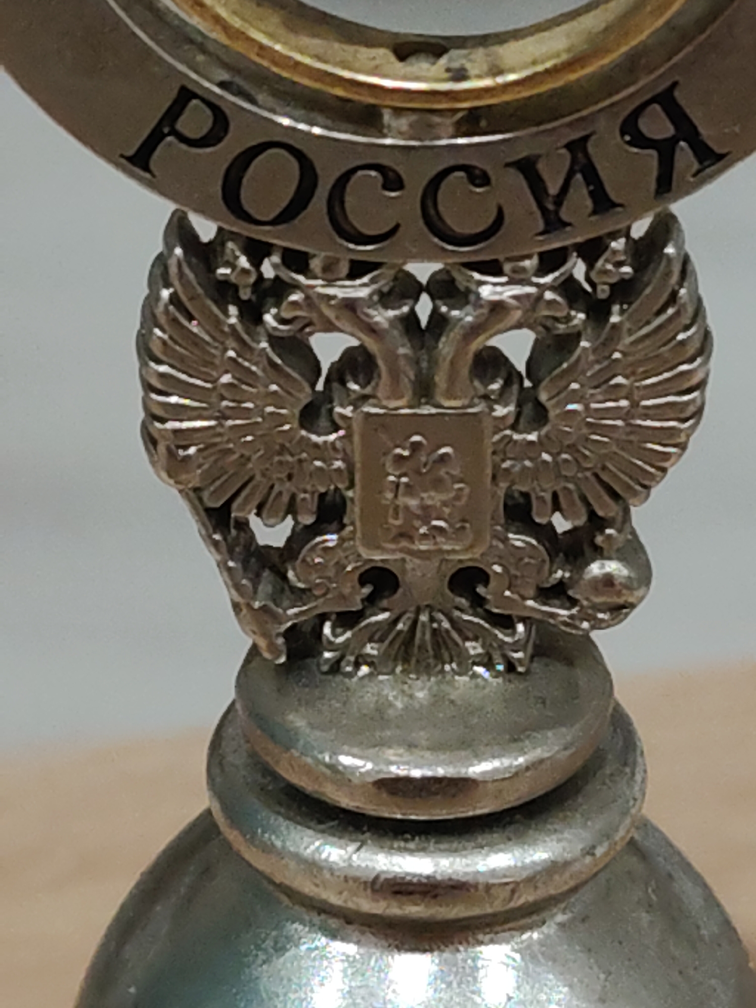 Close-up shot of a silver-colored bell with the word "РОССИЯ" (Russia) inscribed on a circular band near the top. Below the inscription is a detailed depiction of the double-headed eagle, the coat of arms of Russia. The bell has a simple, rounded shape with tiered elements leading up to the eagle. The overall impression is of an ornate, possibly commemorative or decorative object.
