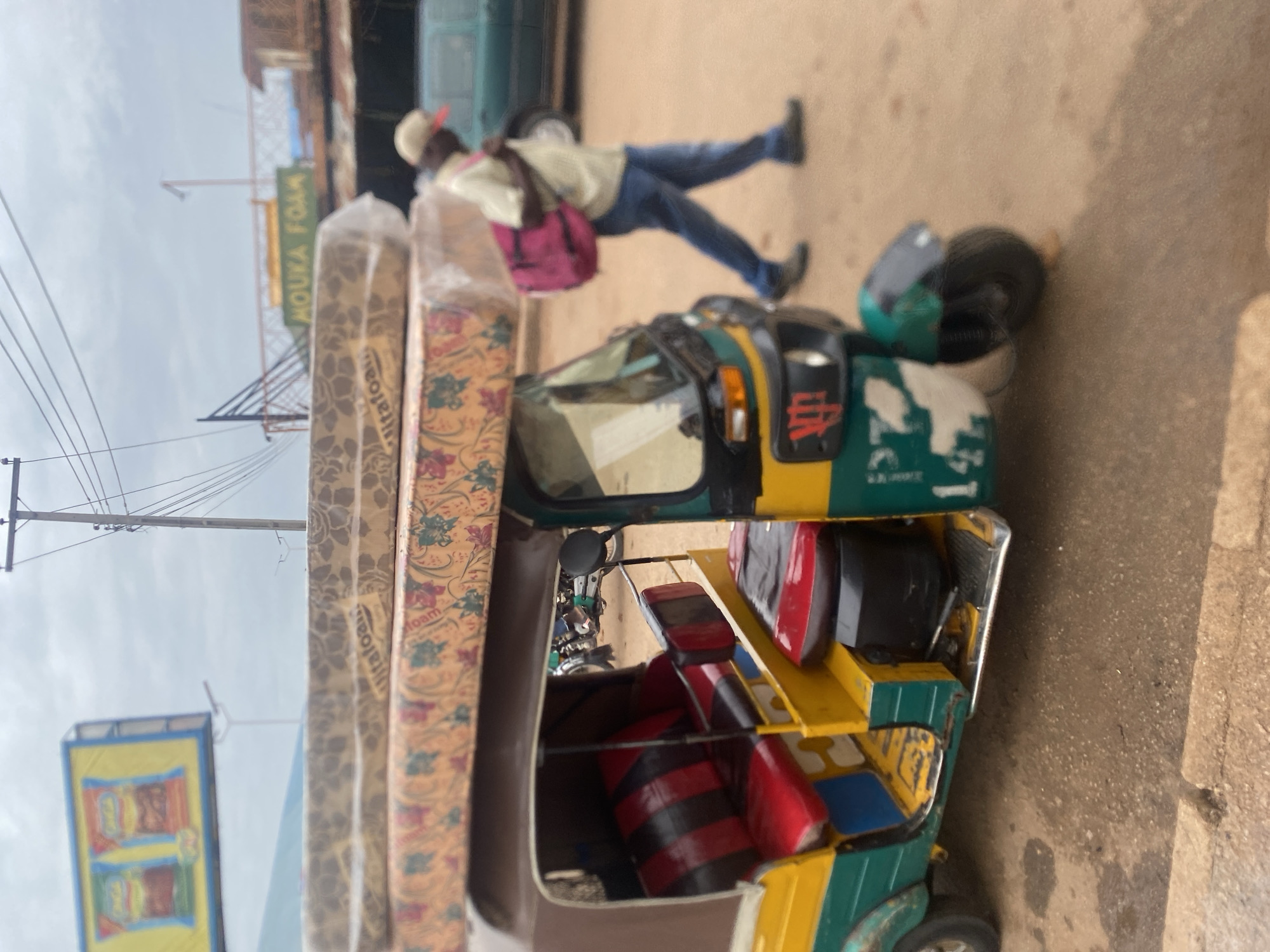 In the city of Bukuru, Nigeria, a three-wheeled auto-rickshaw, painted green and yellow with red and black striped seats, is parked on a dusty road. Two mattresses, wrapped in clear plastic, are stacked on its roof. A man wearing a light-colored shirt, blue jeans, a beige cap, and dark shoes walks away from the auto-rickshaw, carrying a large pink duffel bag over his left shoulder. Behind him, a green vehicle is partially visible. In the background, a billboard displays images of two bags of snacks, and further back, a sign reads "MOUKA FOAM". Power lines and a utility pole are visible against an overcast sky. The ground is a mix of dirt and some wet patches.