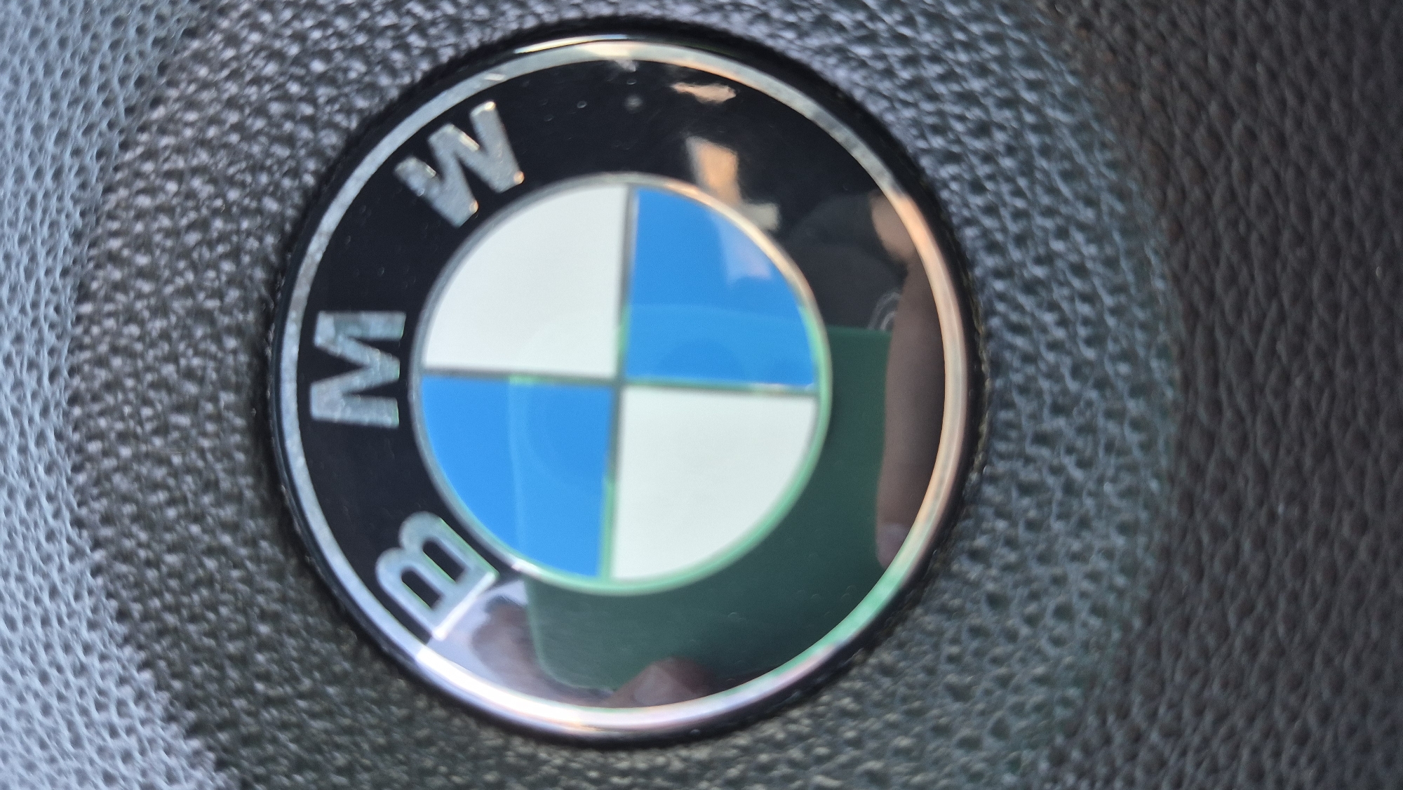 A close-up, slightly out-of-focus image shows the BMW logo on a textured black steering wheel. The logo is circular with a black outer ring containing the letters "B", "M", and "W". Inside the ring, the logo is divided into four quadrants, alternating between blue and white. A reflection of a person holding a phone is visible in the lower left quadrant of the logo. The background is a dark, textured surface, likely the leather of a steering wheel.