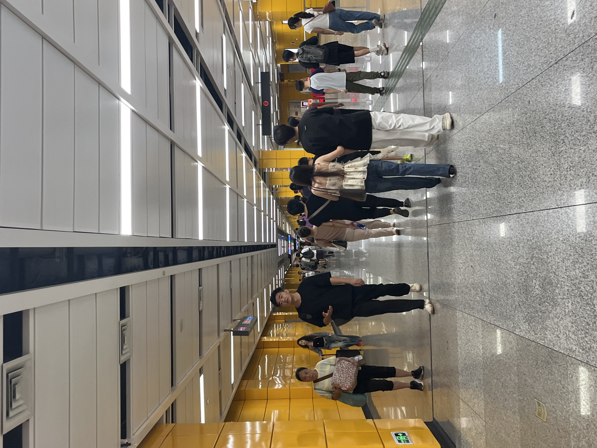 The image depicts a busy underground metro station in Panyu District, China, with a long, brightly lit corridor. The walls are painted a vibrant yellow, and the ceiling features a modern design with recessed lighting and ventilation systems. The floor is a polished gray granite with reflective qualities. Several individuals are walking in both directions along the corridor, some singly and others in small groups.
In the foreground, a woman with a patterned tote bag over her shoulder and a blue bag in her hand walks towards the left. To her right and slightly ahead is a man in a black t-shirt and black pants, looking forward with his left arm slightly raised. Further down the corridor, a group of people are walking away from the camera. One person in this group, a woman, wears a white floral patterned top with blue jeans. To her left, a man in a black t-shirt and white pants also walks away. Behind them, other passengers can be seen, some with backpacks. Traffic flow appears orderly. The overall scene conveys a sense of transit and daily urban life.