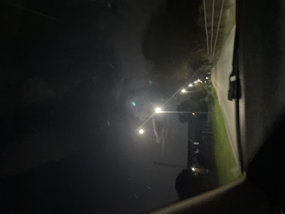 The image captures a nighttime scene from inside a vehicle, looking forward through the windshield, revealing the dashboard and a windshield wiper in the lower foreground. The setting is a wet, paved road in Monsummano Terme, Italy, extending into the distance and illuminated by a series of bright streetlights positioned along its left edge. To the left of the road, a grassy verge precedes a fence, beyond which faint outlines of a low-rise building and a tall, slender industrial structure are visible against the dark sky. The right side of the road features a wooden fence running parallel to the asphalt, backed by dense foliage or trees. The night sky is dark, with scattered clouds partially obscuring a prominent, bright light source, likely the moon. No individuals or specific actions are discernible beyond the implicit presence of the vehicle's occupant.