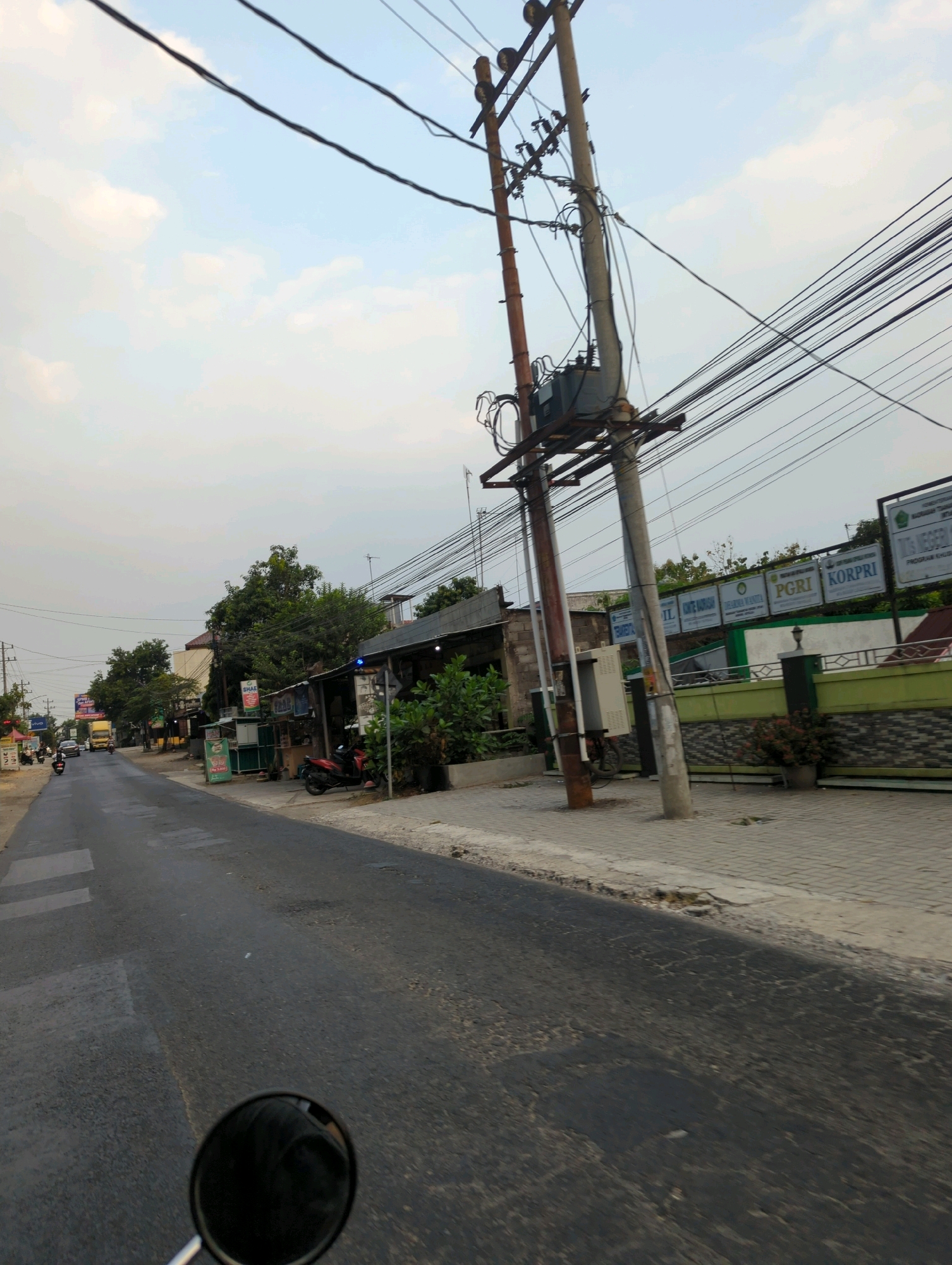 This image depicts a street scene in Kacangan, Indonesia, during what appears to be late afternoon or early evening due to the soft lighting. The perspective is from a moving vehicle, likely a motorcycle, as evidenced by the reflection in the rearview mirror.
The foreground is dominated by the asphalt road, showing signs of wear with patches and cracks. To the right of the road, a paved sidewalk is visible, separated from the road by a curb. Beyond the sidewalk, a green and white striped fence encloses a building or property. Several signs are mounted on this fence, including one that reads "PGRI KORPRI" and another with text in Indonesian, possibly indicating a school or government institution. A utility pole with numerous electrical wires and a transformer stands prominently on the right side of the street.
On the left side of the street, a row of buildings lines the road, some appearing to be shops or small businesses. A yellow truck is visible further down the road, and several motorcycles are parked along the roadside. Lush green trees are interspersed among the buildings, adding to the semi-urban, semi-rural feel of the location. The sky is a pale blue with scattered white clouds. The overall impression is of a quiet, lived-in street in a town or small city.