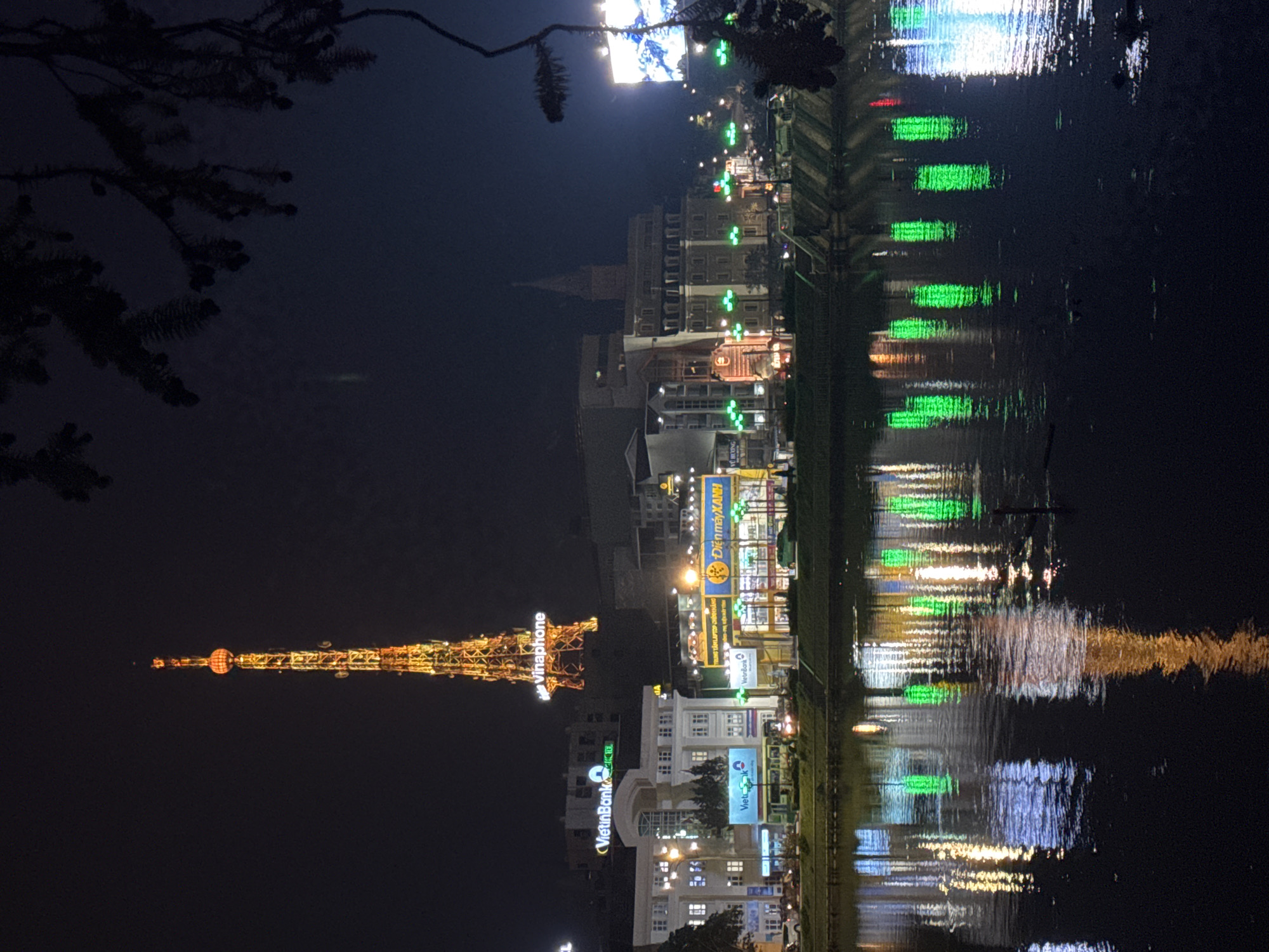 This nighttime image captures a cityscape reflected in dark water. Dominating the left side is a tall, lit structure resembling a transmission tower, adorned with the word "Vinaphone". Adjacent to this, several buildings with illuminated signs such as "VietinBank" and "Điện máy XANH" stand. Further along, toward the center of the image, more buildings are visible, some with a church spire visible at their roofs, with green lights lining the shoreline. The water reflects the lights, creating blurred, elongated shapes of green, white, and yellow, indicating a slight ripple on the water's surface. A dark mass of branches and foliage occupies the top left corner. The scene is likely set in Da Lat, Vietnam, given the presence of the Vinaphone tower, a notable landmark in the city.