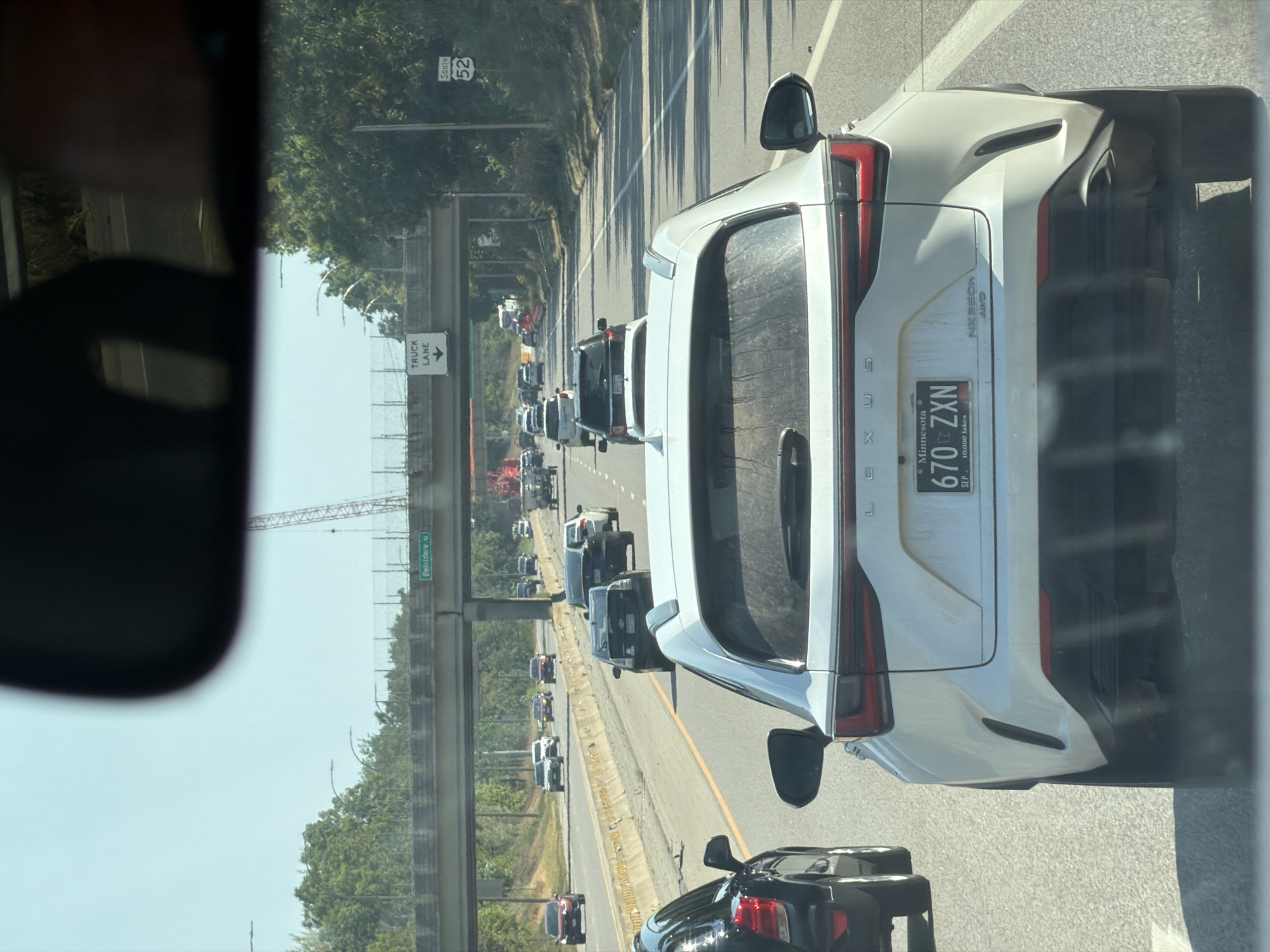 The image captures a view from inside a vehicle traveling on a highway in Saint Paul, Minnesota. The foreground features the rear of a white Lexus SUV with a Minnesota license plate reading "670 ZXN". In the middle ground, multiple vehicles are visible on the highway, including black sedans and SUVs. Overpasses stretch across the highway, with signs indicating "TRUCK LANE" and "SOUTH 52". A construction crane is visible in the distance. The environment appears to be a sunny day with a clear sky.