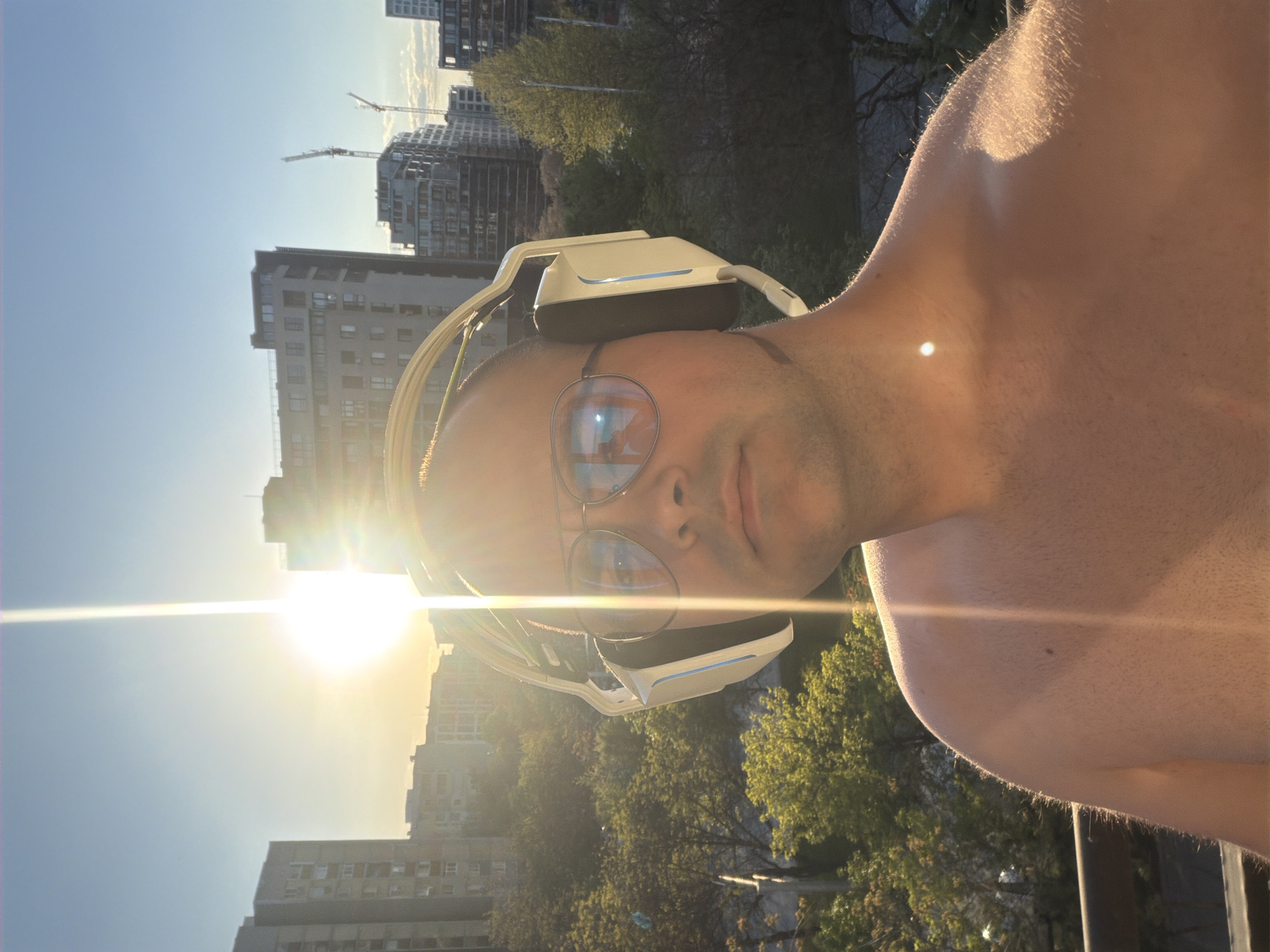 The image, rotated 90 degrees clockwise, presents a close-up portrait of a person outdoors under direct, low-angle sunlight. The subject, likely male, looks directly at the viewer with a neutral expression. They wear circular-framed clear glasses and cream or light-colored over-ear headphones. The sun creates a significant lens flare across the upper right portion of the image, partially obscuring the face but highlighting the headphones and the outline of the head and shoulder. The person's bare shoulder and upper chest are visible, catching the warm sunlight and showing fine body hair.
In the background, an urban landscape unfolds, consistent with the context of Plovdiv, Bulgaria. Several multi-story buildings, some appearing residential or commercial, are visible. Two prominent construction cranes rise above the buildings against the clear blue sky, suggesting ongoing development in the area. Green trees are scattered throughout the mid-ground, adding natural elements to the city scene.
The strong, warm illumination and prominent lens flares indicate the photograph was taken during either sunrise or sunset, often referred to as the golden hour. The sky transitions from a vibrant blue at the top to a lighter, hazy yellow-white near the sun's position. The overall mood is calm, capturing a moment of personal enjoyment in a dynamic city setting. There is no visible text in the image.