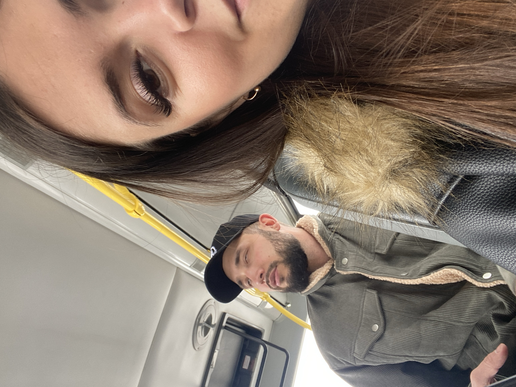 The image shows two individuals, presumably a couple, on what appears to be public transport – potentially a bus or tram. The woman is positioned in the foreground, with the upper portion of her face visible, featuring prominent eyelashes, an earring, and brown hair. She is wearing a jacket with fur trim. Behind her, a man is seen wearing a cap and a corduroy jacket with shearling trim. He has a beard and his eyes are closed, suggesting he may be sleeping. Yellow handrails are visible in the background, along with the interior of the vehicle, which includes a door.
Given the context provided, there is no visible evidence or definitive information to ascertain the location is in the city of Chaikovsky, Russia. If the location is in that area, it’s most likely that this is a daily occurrence on public transport.