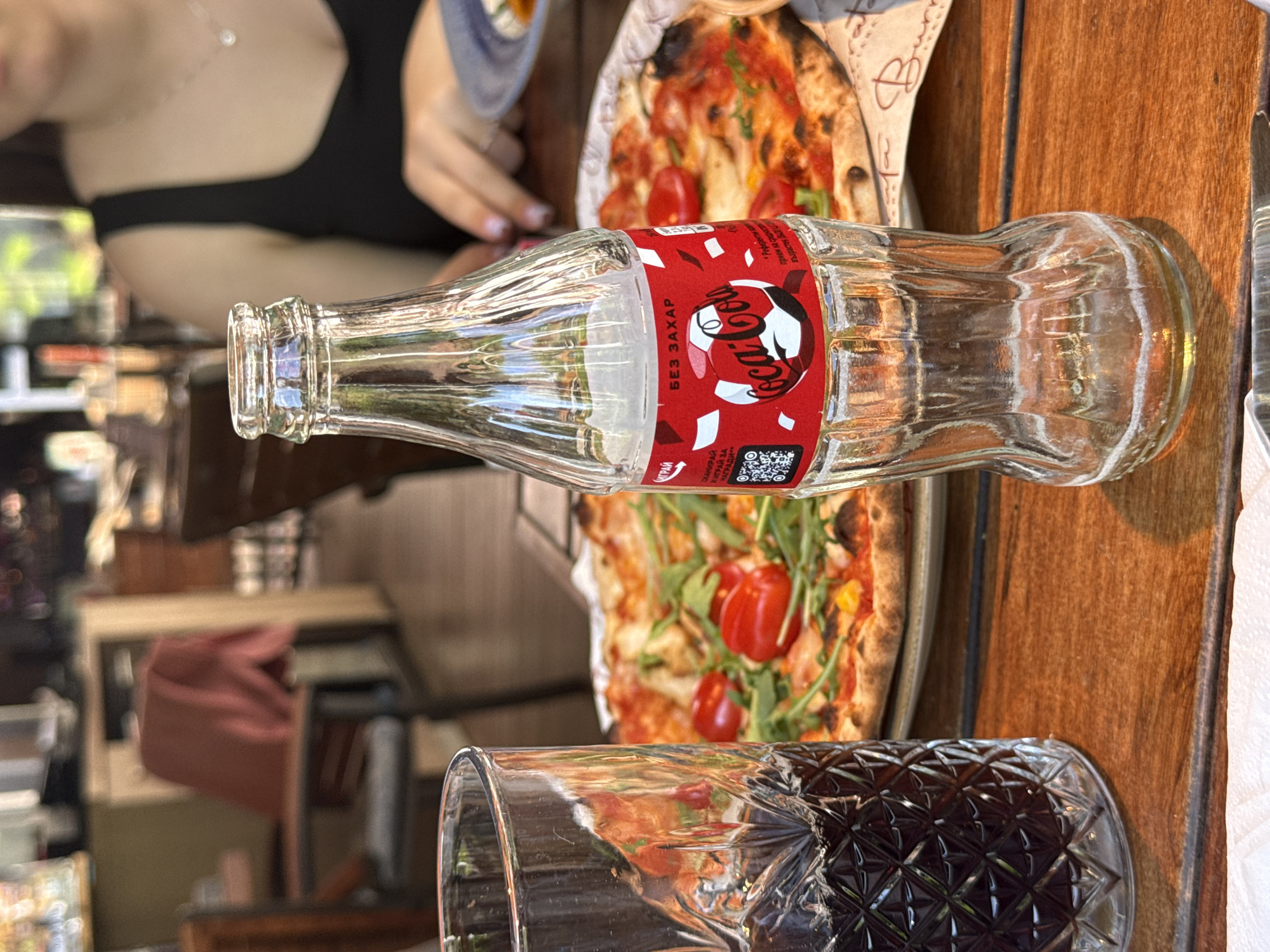 A close-up, eye-level shot shows a partially empty glass bottle of Coca-Cola Zero Sugar standing upright on a wooden table. The bottle has a red label with the Coca-Cola logo and text. To the left of the bottle is a tall glass filled with a dark beverage, likely cola, with ice. In the background, a pizza with cherry tomatoes and arugula is visible on a plate. A person's shoulder and chest wearing a black tank top and a necklace are out of focus in the upper right corner of the image. The background suggests an outdoor seating area of a restaurant with tables, chairs, and foliage. The scene is likely set in Sofia, Bulgaria, as indicated by the Coca-Cola Zero Sugar bottle label which is in Bulgarian Cyrillic.