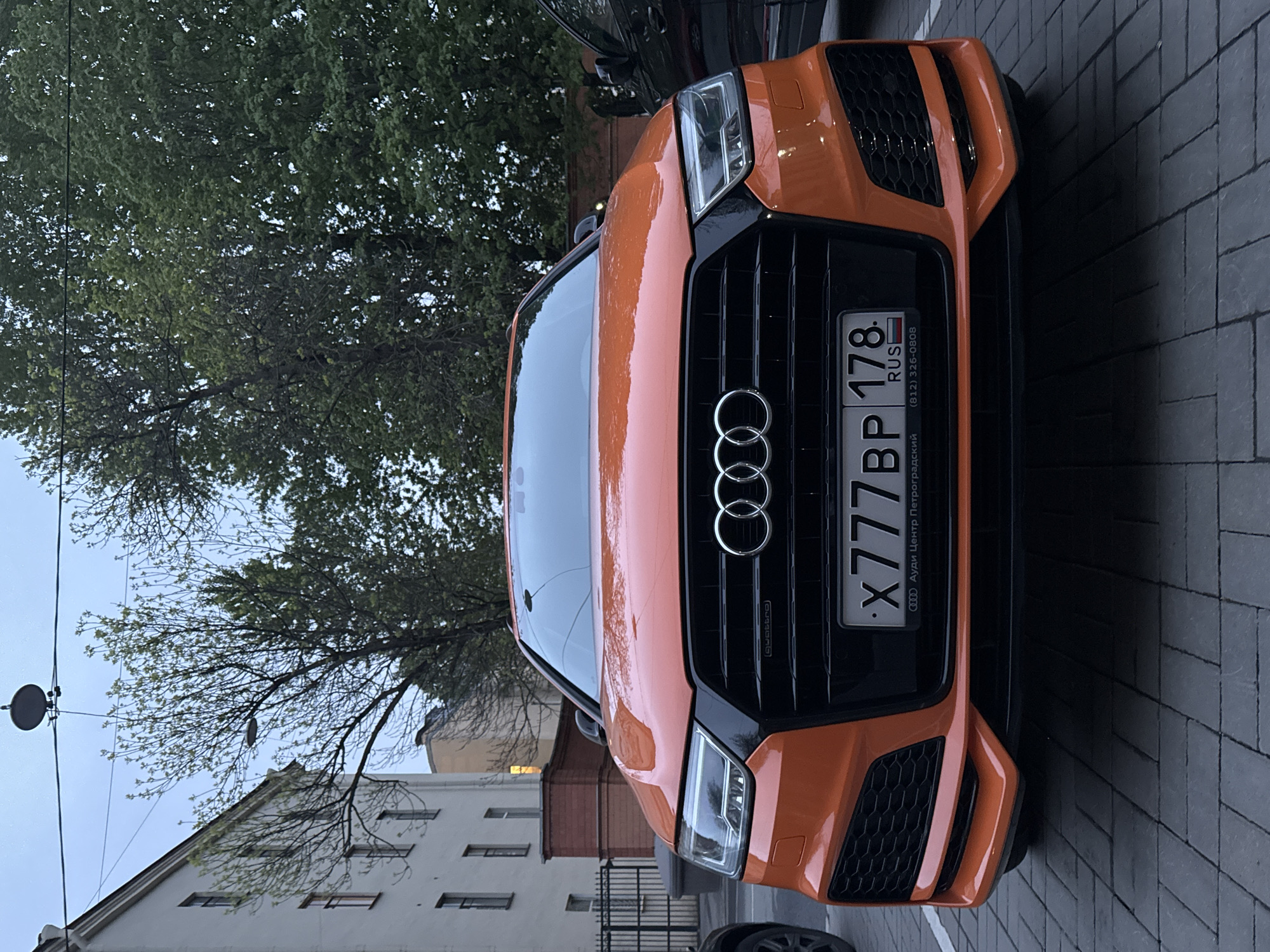 The image shows the front of an orange Audi car, likely an SQ5 model, parked on a cobblestone street. The car has a Russian license plate reading "X777BP 178". The Audi logo and "quattro" badge are visible on the grille. The license plate indicates the car is registered in Saint Petersburg, Russia. The text on the license plate frame is in Russian and states "Audi Center Petrogradsky," along with a phone number. In the background, there are buildings with white facades and trees with green leaves, typical of a European city street. The sky is overcast, suggesting it may be early morning or late afternoon. Another vehicle, possibly dark-colored, is partially visible at the top of the frame.