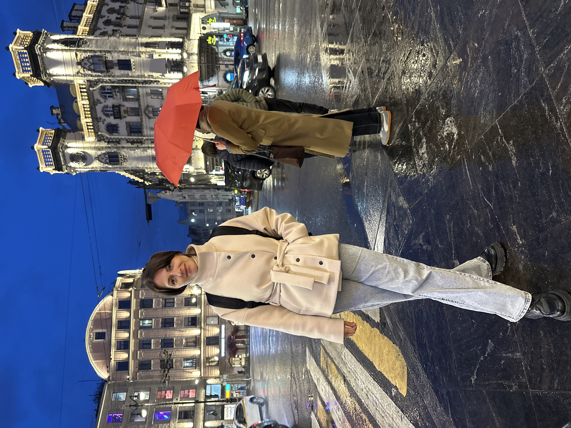 The image shows a night scene in Saint Petersburg, Russia, likely during or after rain, as the ground is wet and reflective. A young woman with short brown hair stands in the foreground, facing the camera with a slight smile. She is wearing a beige belted jacket, light wash jeans, and black boots, with a black backpack. Behind her, across the street, is the illuminated architectural facade of Au Pont Rouge (sometimes referred to as the "House at the Red Bridge"). To the right, people are walking with an orange umbrella and a trench coat. The sky is a deep blue, indicating evening. Several cars are visible, their headlights adding to the illumination. The wet pavement is likely made of black stone slabs.