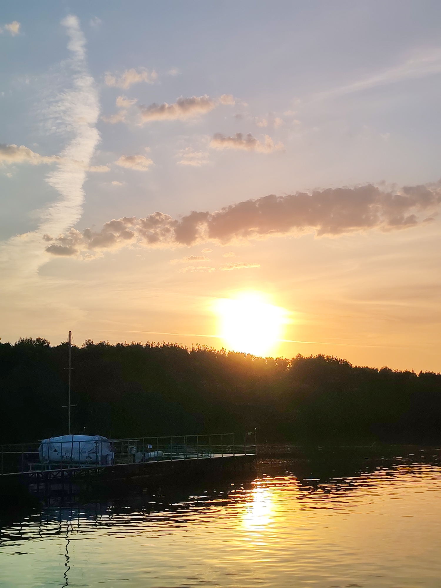 The image depicts a sunset over a body of water, possibly a lake or river. In the foreground, there's a dock with a white covered boat and railing. The water reflects the golden light of the setting sun. The middle ground features a line of trees silhouetted against the bright sky. The sky is a mix of light blues, yellows, and oranges, with clouds. Based on the query, the location is in Ermolino, Russia. The scene lacks any people or direct actions.