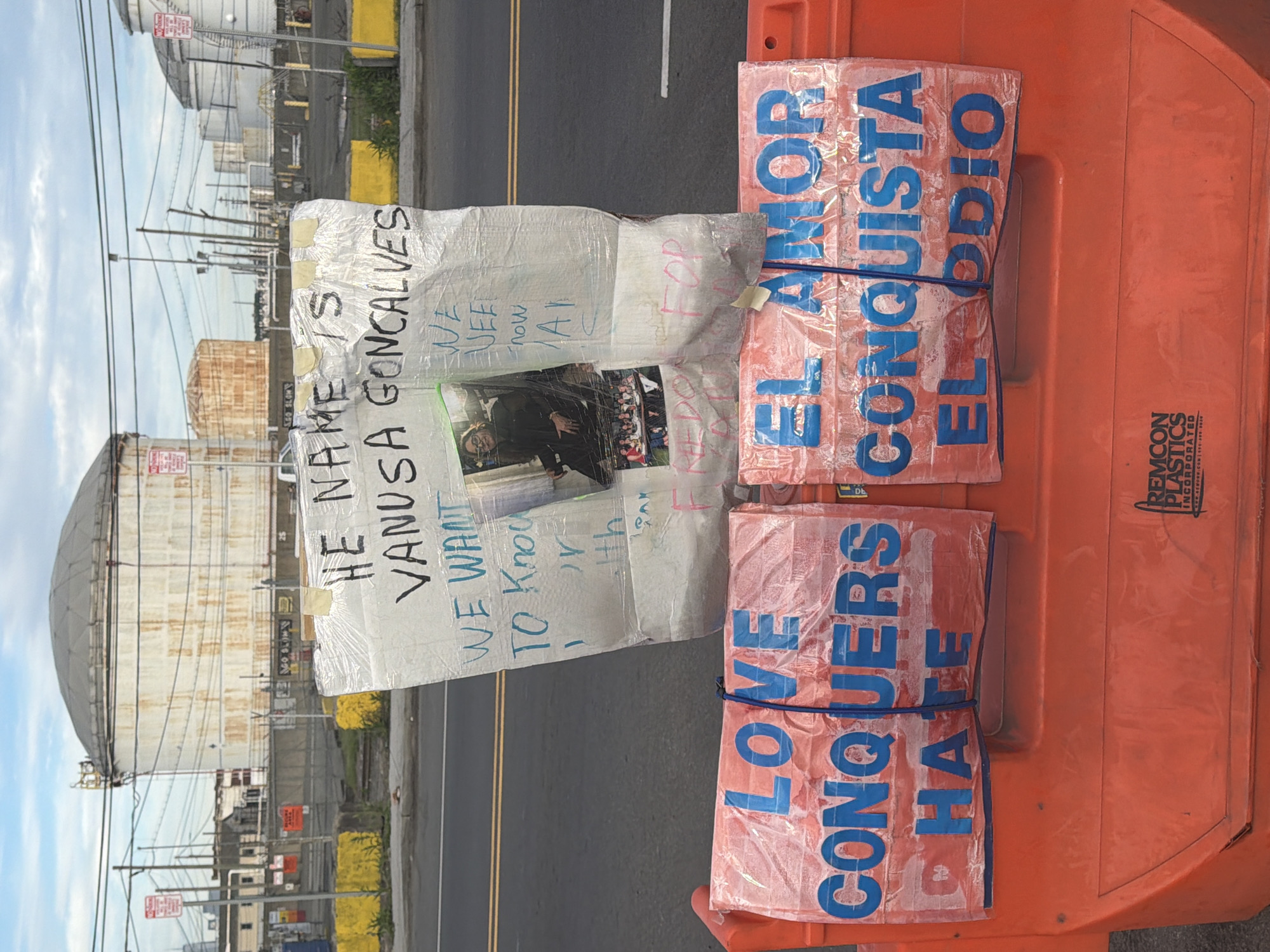 The image shows a sign attached to an orange plastic barrier on a city street. The sign appears to be homemade and contains a photo and handwritten text reading "HE NAME IS VANUSA GONCALVES, WE WANT TO KNOW NOW". Another attached sign has "LOVE CONQUERS HATE" and "EL AMOR CONQUISTA EL ODIO" printed on it. The background shows oil/gas storage tanks. Based on the oil tanks and streetscape the location can be identified as Newark, United States of America.