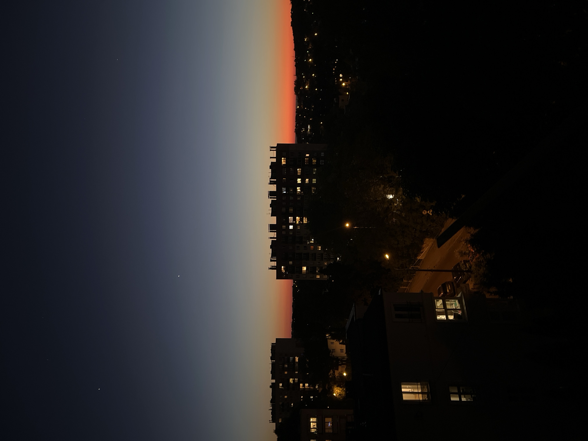 The image depicts an urban cityscape at dusk or night, oriented vertically. The sky transitions from a dark blue on the left to a gradient of gray and orange near the horizon. Buildings with illuminated windows are visible, suggesting residential occupancy. Streetlights punctuate the scene along a road. Silhouetted trees and vegetation are interspersed among the buildings, adding depth to the composition. The location appears to be in an unknown city. No people are visible.