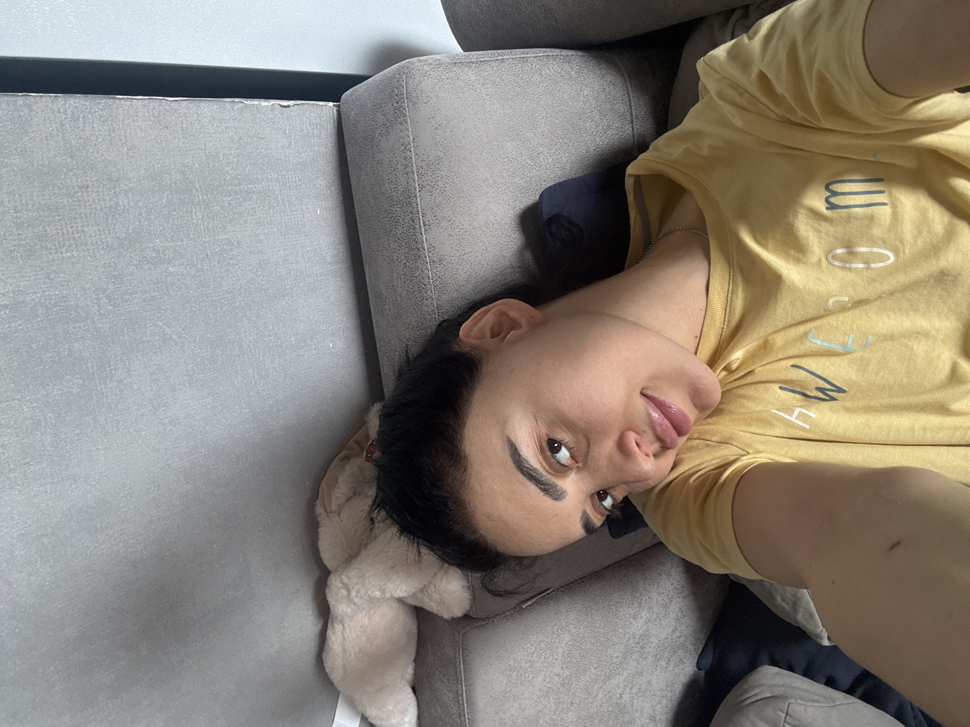 A woman lies on a gray sofa next to a stuffed bunny toy. She wears a yellow t-shirt. The background is a gray wall. The location cannot be determined from the image.