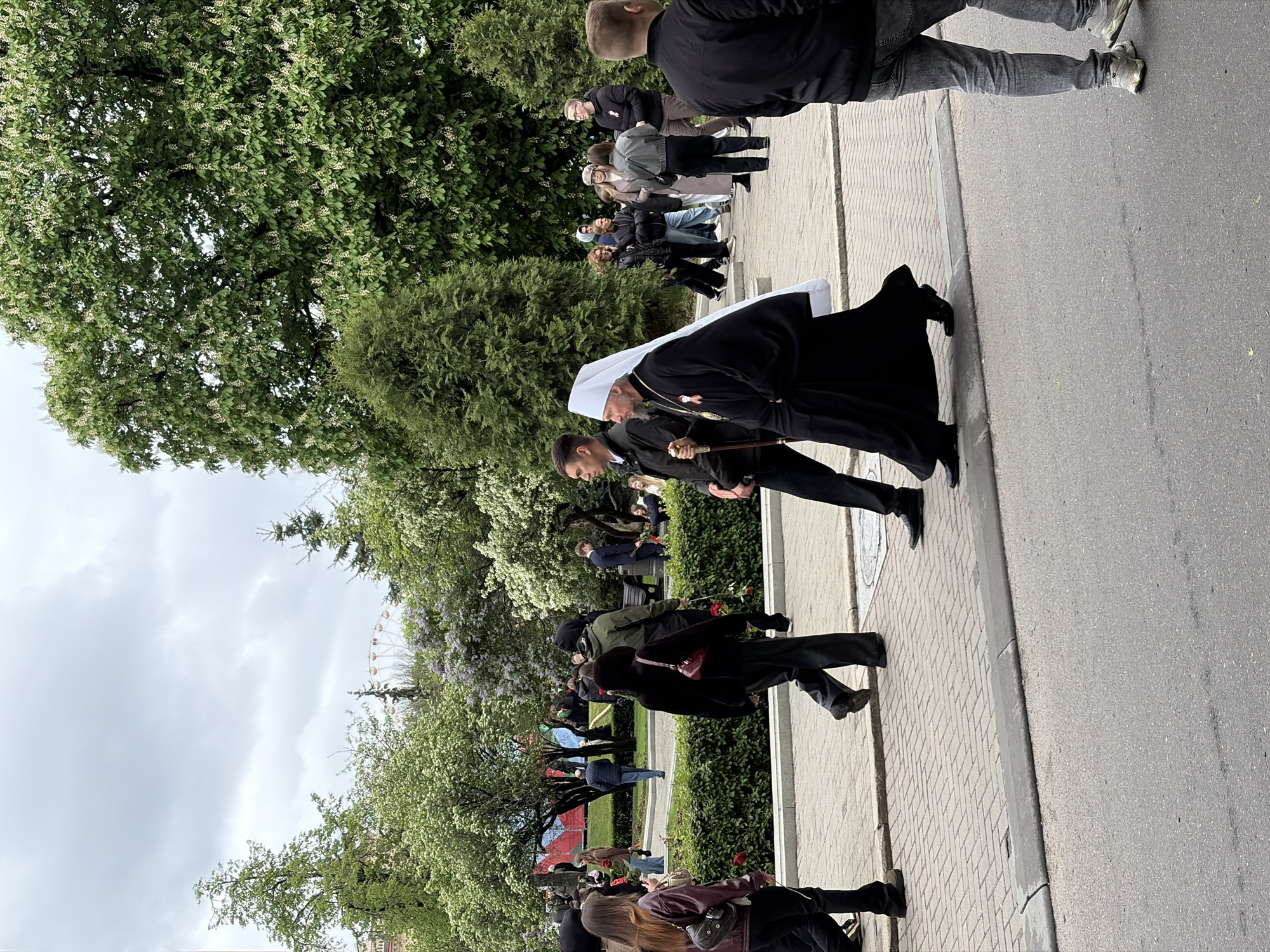 The image shows a procession along a brick-paved sidewalk bordered by a street and a park area with trees. The main subject is a man in an elaborate black robe and a white headdress, likely a religious figure, walking with the aid of a person holding a decorative staff. A line of people stands along the sidewalk, appearing to observe or participate in the procession. In the background, a Ferris wheel is visible, suggesting the location is an urban park area.