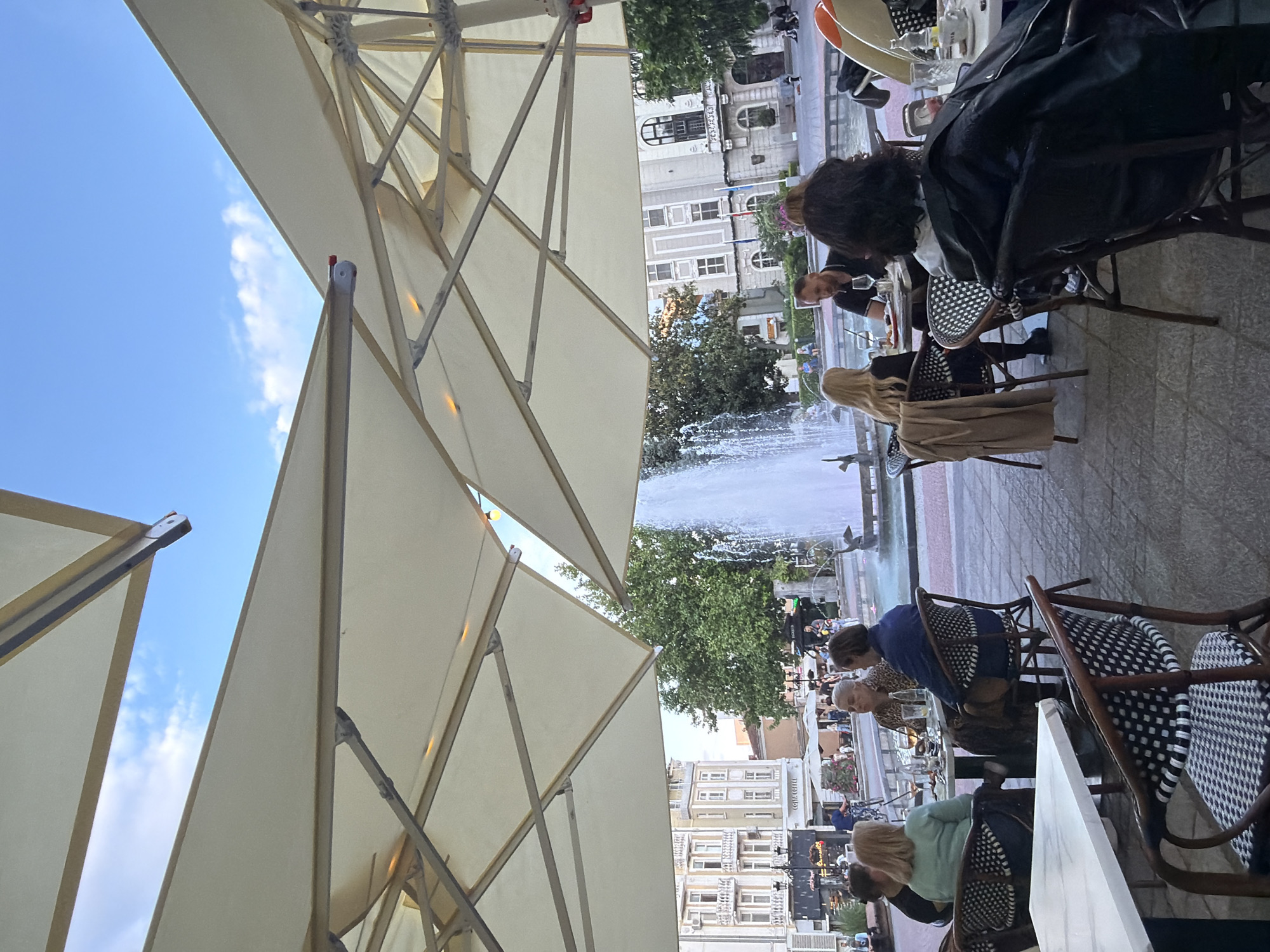 The image captures an outdoor dining scene in Plovdiv, Bulgaria, near a prominent fountain. The viewpoint is from under large, light-colored parasols, looking out at the square. Several people are seated at tables, some appearing to be eating or drinking. A fountain with shooting jets of water is visible in the background, with trees and buildings beyond. Some individuals are interacting at their tables. The setting is a paved pedestrian area typical of a city square.