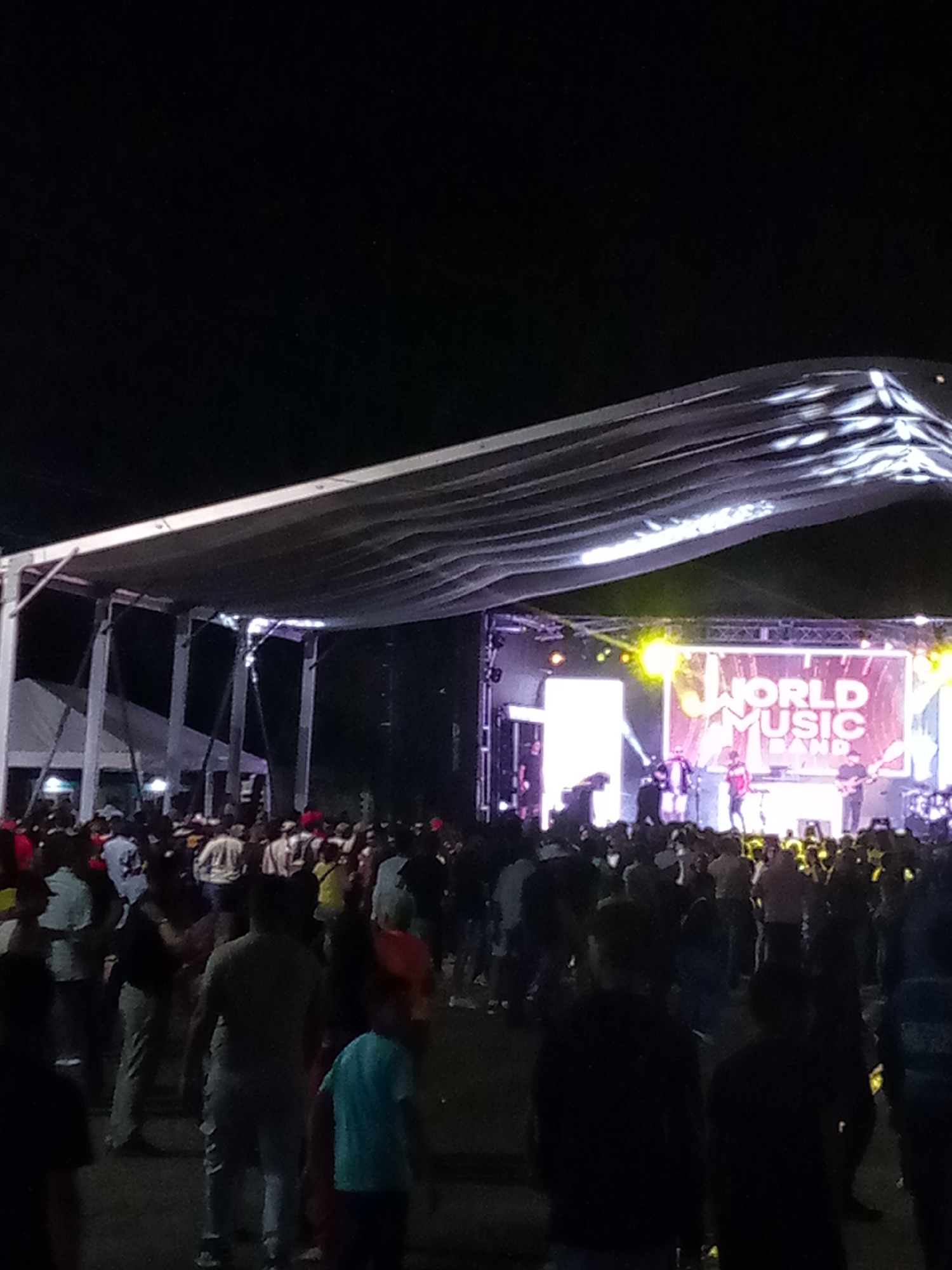 The image shows an outdoor night concert in San Juan Bautista, Venezuela. A crowd of people is gathered in front of a stage where a band is performing. The stage is lit up with colorful lights, and a large screen displays "WORLD MUSIC BAND." The audience is a mix of adults and children, dressed in casual clothing. Some people are standing and dancing, while others are sitting or leaning against the stage. The atmosphere appears festive and energetic.