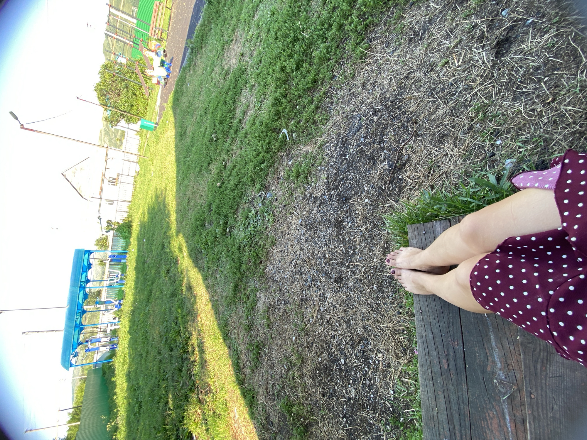 The image depicts an outdoor scene in Maloarslangulovo, Russia, likely a public park or recreational area. In the foreground, a person's bare feet and legs, clad in a maroon polka-dot dress, are visible resting on a wooden bench. The feet show dark red nail polish. The ground in front of the bench is a mix of dry grass and gravel. In the middle ground, a blue-covered outdoor gym structure with various exercise equipment is present, surrounded by green grass. Further in the background, there's a white building with a pitched roof, a green metal fence, and trees. To the right, a playground with a colorful see-saw, a bench, and part of a slide structure is visible. The sky is bright and appears to be a clear day.