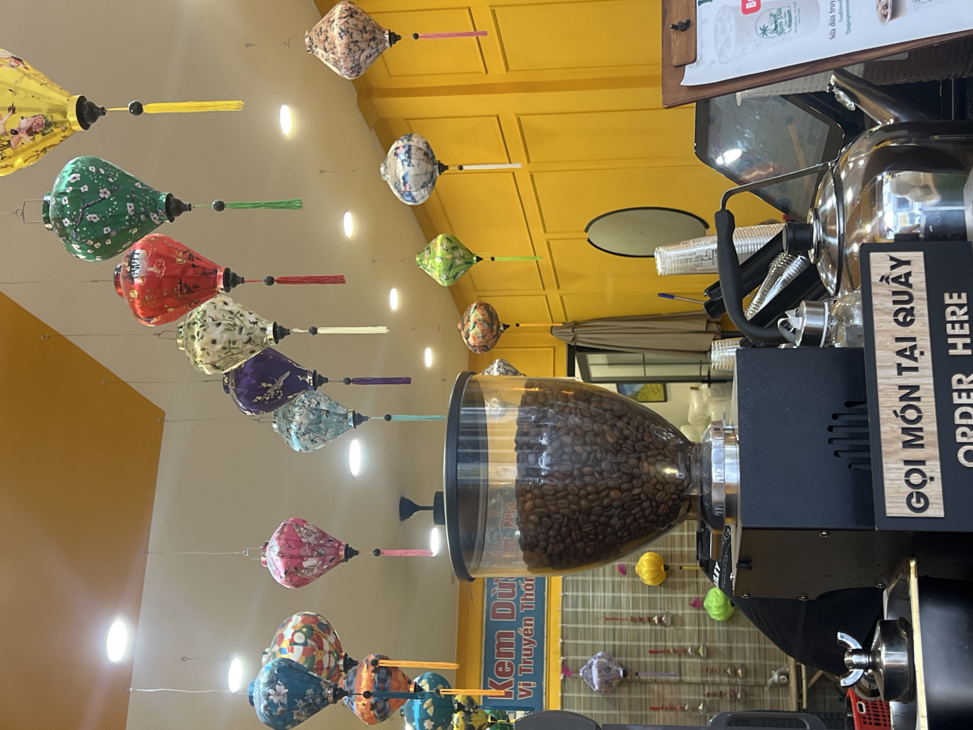 The image shows the interior of a cafe in Nha Trang, Vietnam, decorated with numerous colorful Vietnamese lanterns hanging from the ceiling. In the foreground, a large, transparent coffee bean grinder is prominent, filled with roasted coffee beans. To the right of the grinder, a metal teapot sits on a counter, alongside other cafe equipment such as disposable cups and a tablet device. A menu board with Vietnamese text is also visible on the right. In the background, a yellow wall is decorated with a grid pattern, and a striped bamboo mat is visible behind the grinder. The overall atmosphere is vibrant and inviting, typical of a local establishment in the city.