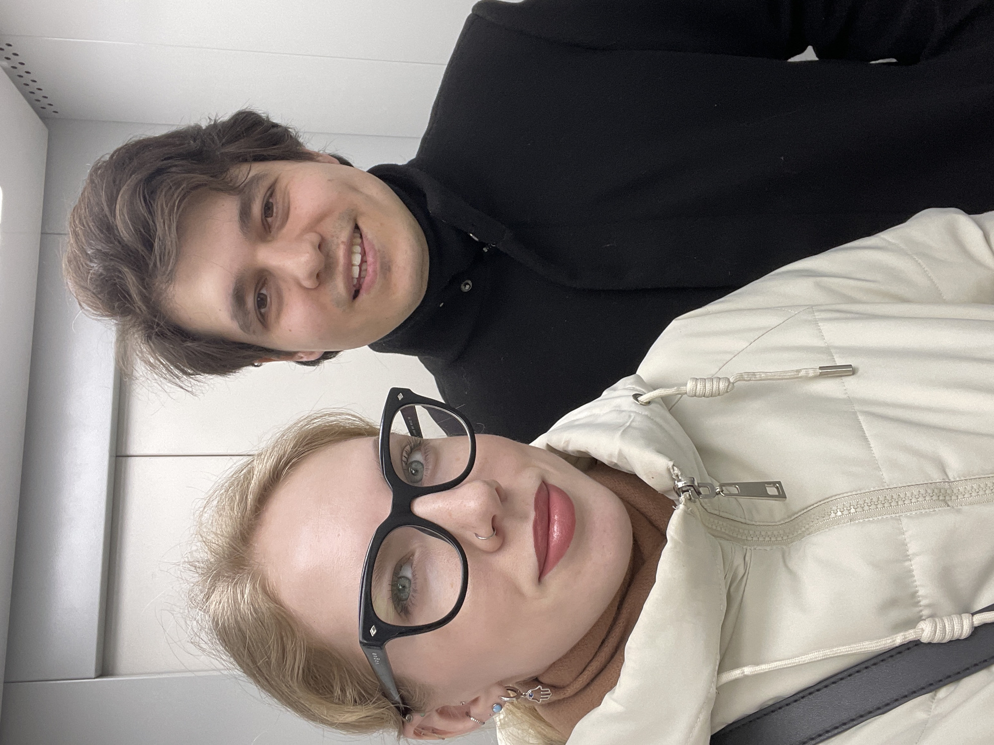 Two people are pictured indoors. A man with brown hair, wearing a black turtleneck or coat, is visible in the upper-left portion of the frame, smiling. A woman with blonde hair wearing black glasses and a light-colored puffy jacket is positioned closer to the camera in the lower-right portion of the frame. The background appears to be a plain, light-colored wall, possibly within an elevator or similar enclosed space. There is no evidence to suggest this is in Izhevsk, Russia.