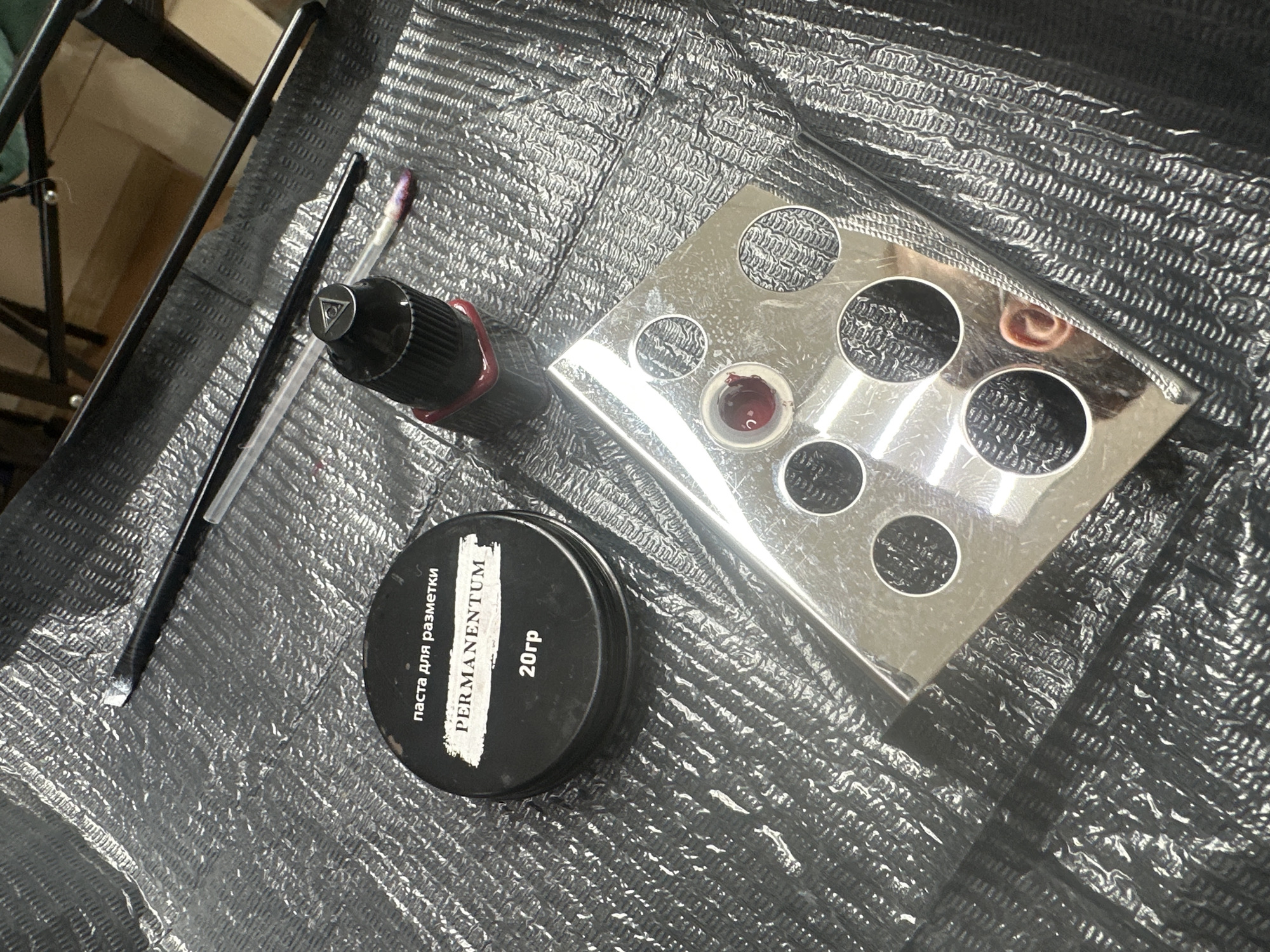 The image depicts a setup for permanent makeup or tattooing, likely in Chaikovsky, Russia. A black, textured surface, likely a disposable sheet, forms the background. From left to right are a couple of fine-tipped brushes, a permanent makeup machine with a cartridge on it, a black jar labeled "PERMANENTUM" in Cyrillic with the text "Паста для разметки" (paste for marking) and "20гр" (20g), and a shiny metal palette with circular depressions, one of which contains a small amount of red pigment. The scene is lit well.