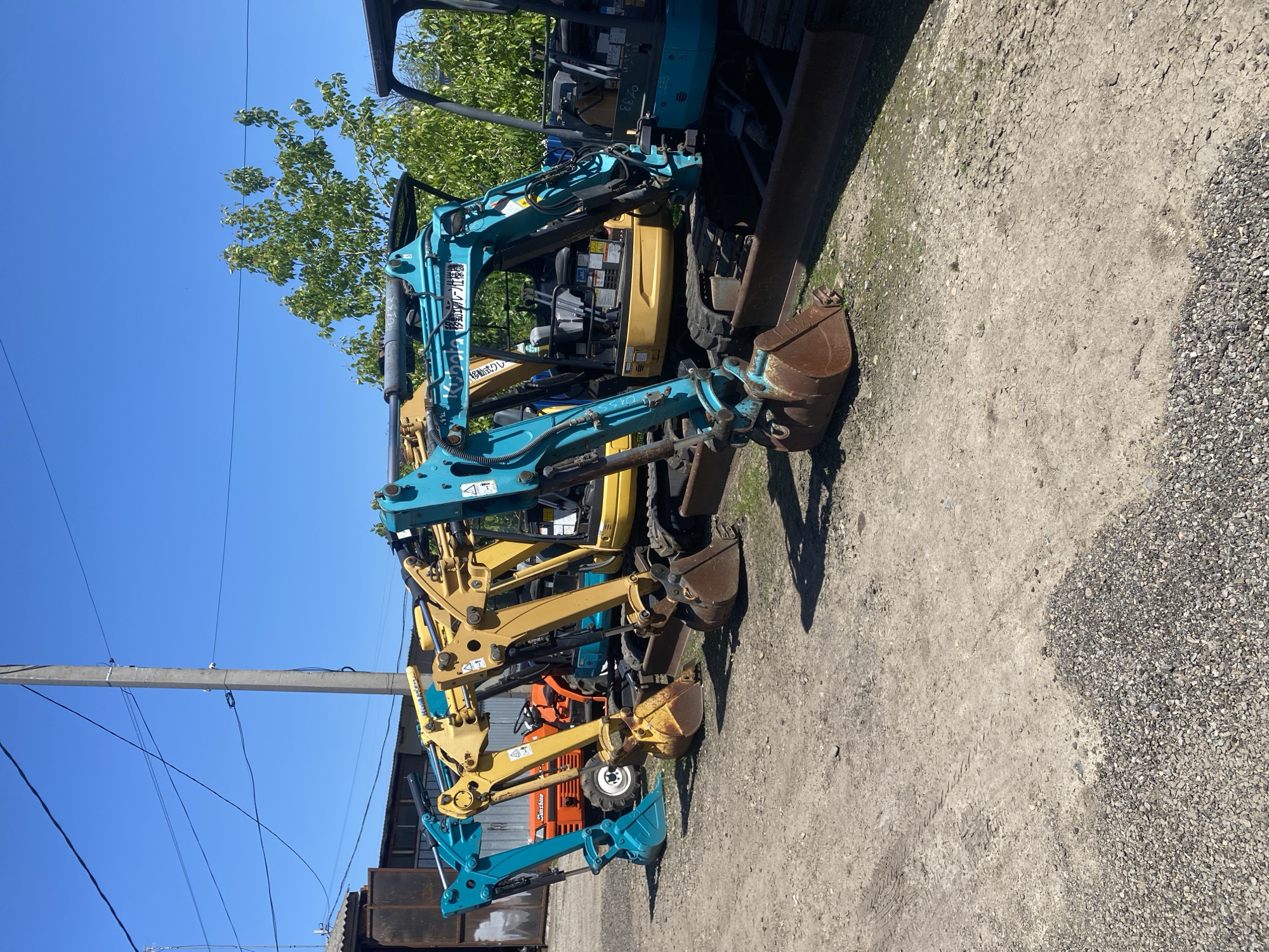 The image shows a group of small excavators parked on a gravel and dirt surface. The excavators are various colors, including blue, yellow, and orange. A utility pole is visible on the left side of the image, with overhead power lines. A tree is visible in the background. The scene is located outdoors on a clear day. The location is not verifiable as Neftyanik, Russia from the image. There are no people visible in the image.