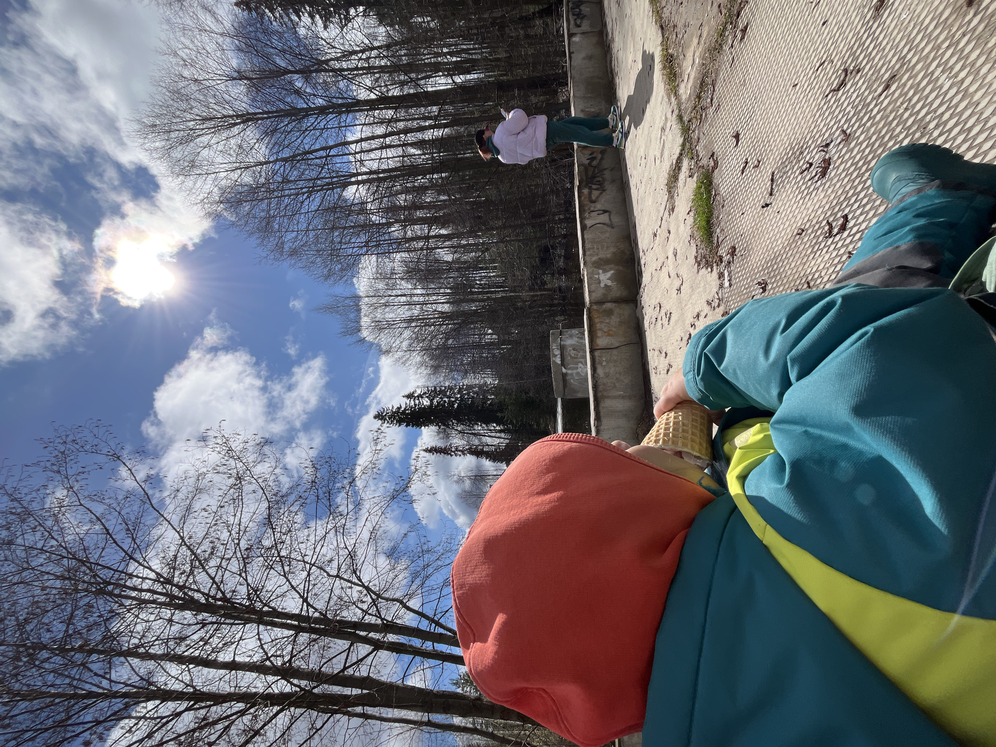 An eye-level shot captures a person holding an ice cream cone in their right hand, with the camera looking upward towards the sky. The individual wears an orange hooded sweatshirt and a teal jacket with a yellow stripe. The ice cream cone is made of a waffle material and appears to contain ice cream. The shot is taken on a sunny day with a bright blue sky and scattered white clouds. In the background, a forest of bare trees is visible, and to the right, a second person can be seen standing on a concrete surface. The location is Chaikovsky, Russia.