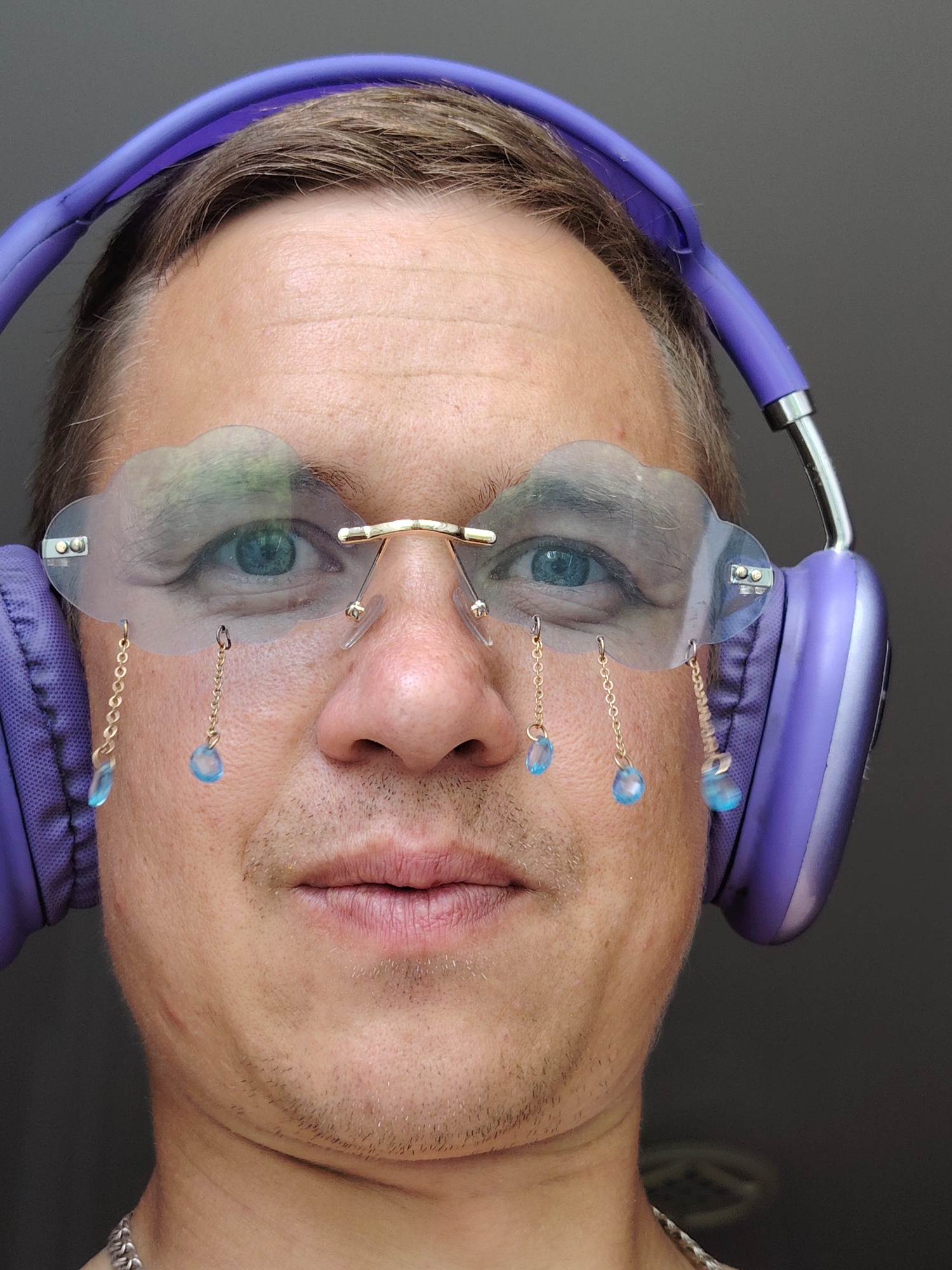 A close-up, waist-up shot features a fair-skinned person wearing large, purple over-ear headphones. The person has short, brown hair and is looking directly at the camera with blue eyes. They are wearing unique, clear glasses shaped like clouds, with gold frames. Dangling from the glasses are gold chains with light blue, teardrop-shaped charms, resembling raindrops. The person has stubble on their chin and upper lip. A silver chain necklace is visible around their neck. The background is a plain, dark gray wall. The scene is indoors, with no indication of the city of Chaikovsky, Russia.