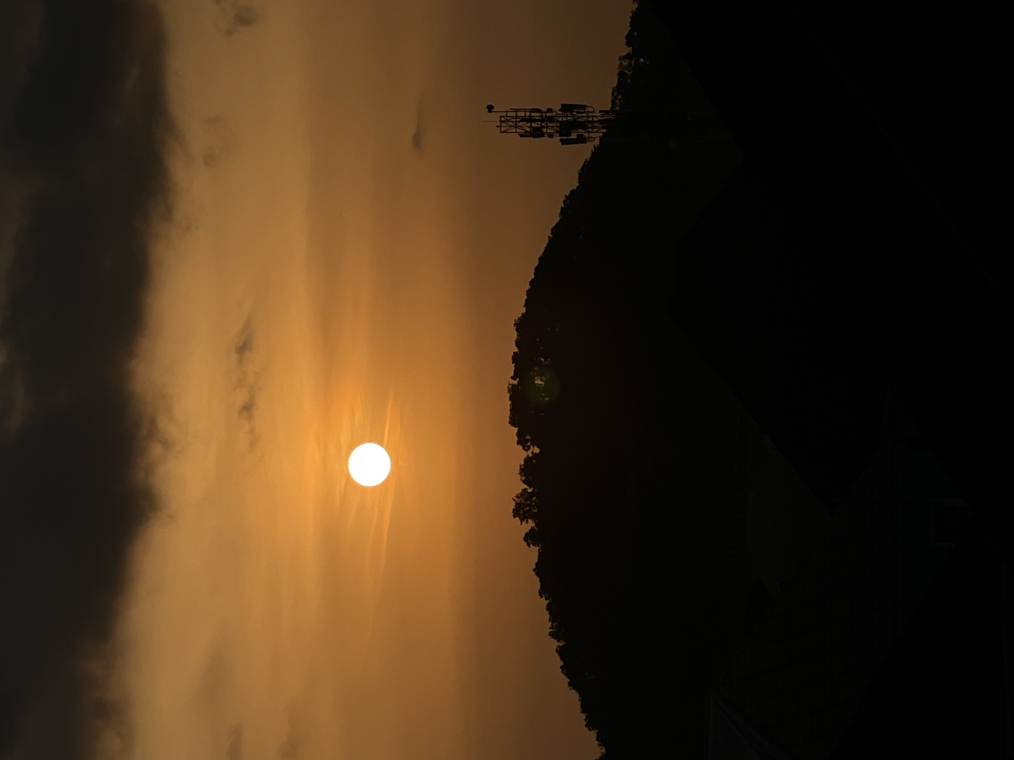 A dusk scene captures the sun, a bright orb against an orange sky, partially obscured by dark clouds. The sun is positioned in the upper center of the frame. Below the sky, a silhouetted mountain ridge spans the mid-ground, its crest outlined with dark trees. To the right of the mountain, a telecommunications tower stands tall, also silhouetted. In the foreground, the dark shapes of rooftops suggest a residential area. The overall lighting is dim, typical of sunset. The location is identified as Abuja, Nigeria. There are no people or animals visible in the image.