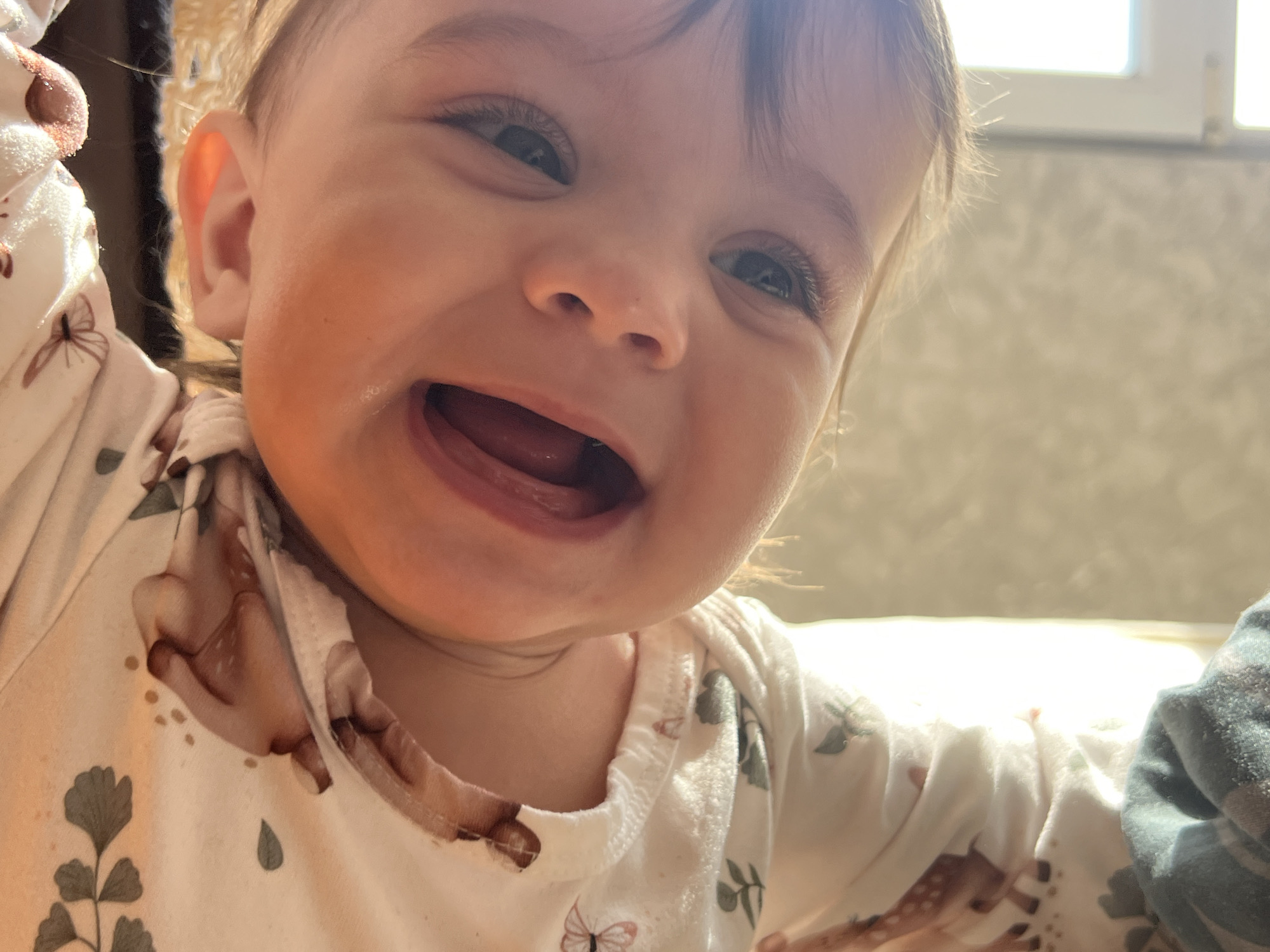 A close-up, high-angle image depicts a young child, appearing to be a baby or toddler, centered in the frame. The child has fair skin, light brown hair, and wide-open eyes directed towards the camera. The mouth is open in a broad smile, revealing gums and the tongue, with small droplets of liquid visible on the chin and around the lower lip. The child wears a light-colored, long-sleeved garment, possibly a onesie, patterned with brown butterflies, green foliage, and brown deer motifs. Portions of the child's arms are visible at the sides, raised slightly. The background is an out-of-focus indoor setting, with a bright, rectangular light source, consistent with a window, visible on the upper right, illuminating the scene with natural light. A blurry, light-colored wall or surface is present behind the child. The image provides no discernible visual markers to confirm a specific location within the city of Elena, Bulgaria.