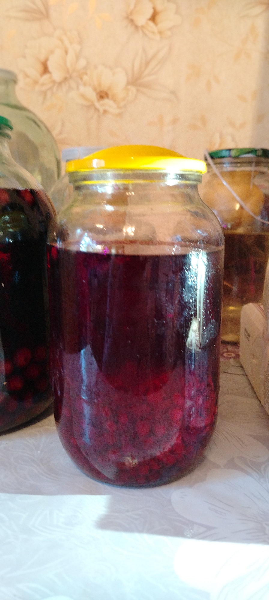 The image shows three glass jars filled with liquid and fruit, likely compote. The jar in the center is largest, filled with a dark red liquid and red berries. It has a yellow lid. To the left is a smaller jar with a green lid, also filled with dark red liquid and red berries. To the right is a jar with a brown liquid. All the jars are on a white surface with a floral pattern. The background is a wallpaper with large floral patterns. The image was taken in Зипуново, Russia.