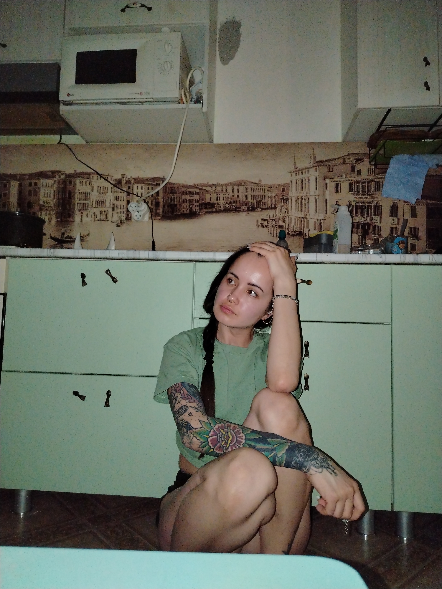 A young woman with a long dark braid and arm tattoos sits on the floor in a kitchen. The kitchen features light green cabinets with antique-style pulls, and a microwave mounted above the counter. A mural depicting a cityscape is visible behind the counter, which holds various household items. The woman is wearing a light green t-shirt. Her expression is pensive as she rests her hand on her head.