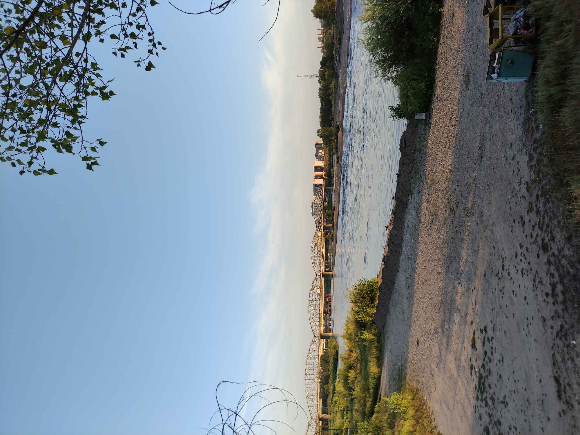 The image captures a wide-angle view of the Irtysh River in Semey, Kazakhstan, during the late afternoon or early evening. A large, multi-span truss bridge, likely the Semey Bridge, dominates the middle ground, stretching across the river and connecting to the city on the far bank. On the far bank, buildings of varying heights are visible, including what appear to be apartment blocks and potentially a church with a dome. A tall radio tower stands out against the sky.
The foreground consists of a sandy embankment leading down to the water. Vegetation, including grasses and bushes, grows along the riverbank and on the embankment. In the lower right corner of the foreground, two industrial-style waste bins are situated, one green and one yellow, both containing visible refuse.
The sky is clear and blue with a few wispy clouds. The sun is low, casting long shadows across the sandy ground and illuminating the bridge from the side, giving it a warm, golden hue. Some bare, thin branches of a tree are visible in the upper left and right corners, framing the scene. The river itself appears calm with gentle ripples reflecting the sky. Several small birds are scattered along the riverbank in the middle distance.