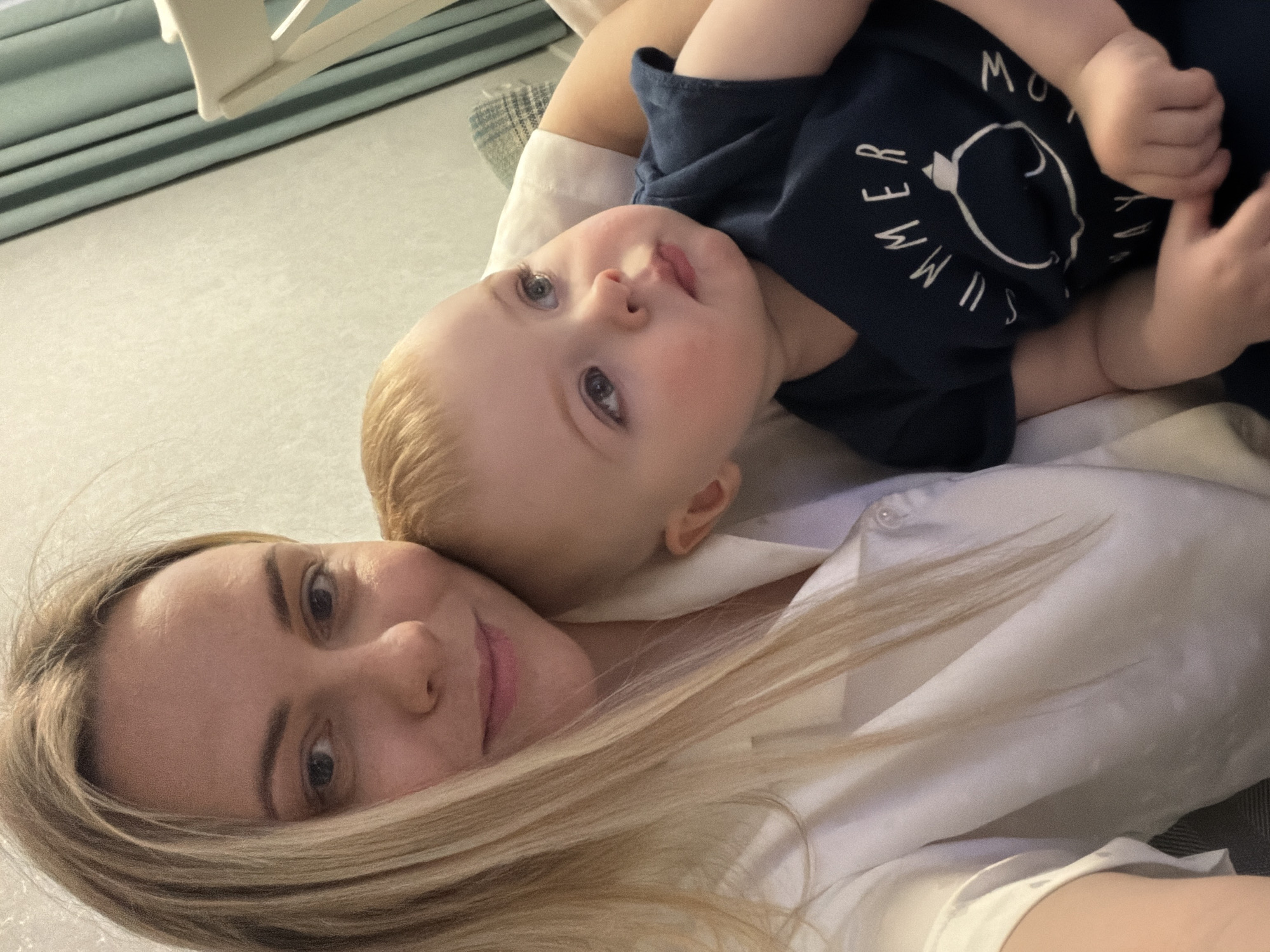 This media file captures a tender indoor moment between a woman and a baby, likely a mother and child, presented from a selfie-style perspective. The woman, positioned in the lower left of the frame, has long, light blonde hair that cascades around her face. She is wearing a white collared shirt and looks directly at the camera with a calm expression and a slight, gentle smile.
Resting on the woman's chest or shoulder, and facing the camera, is a baby or toddler with fair skin and light blonde hair. The child's wide, light-colored eyes convey a sense of innocent curiosity as they look towards the viewer. The baby is dressed in a dark blue t-shirt that features white text and a graphic design. Visible text on the shirt includes "SUMMER" vertically along the left side and "MOW" above a circular graphic on the right, forming what appears to be "MOW SUMMER". Small, chubby hands are also visible near the child's torso.
The background primarily consists of a bright, light-colored surface, suggesting they are lying on a bed or sofa. In the upper left quadrant of the image, hints of light blue-green striped curtains or blinds and a cream-colored furniture piece are discernible. The lighting is soft and even, contributing to the peaceful and affectionate mood of the scene. The image beautifully conveys the close bond between the two subjects in a casual, relaxed setting.
